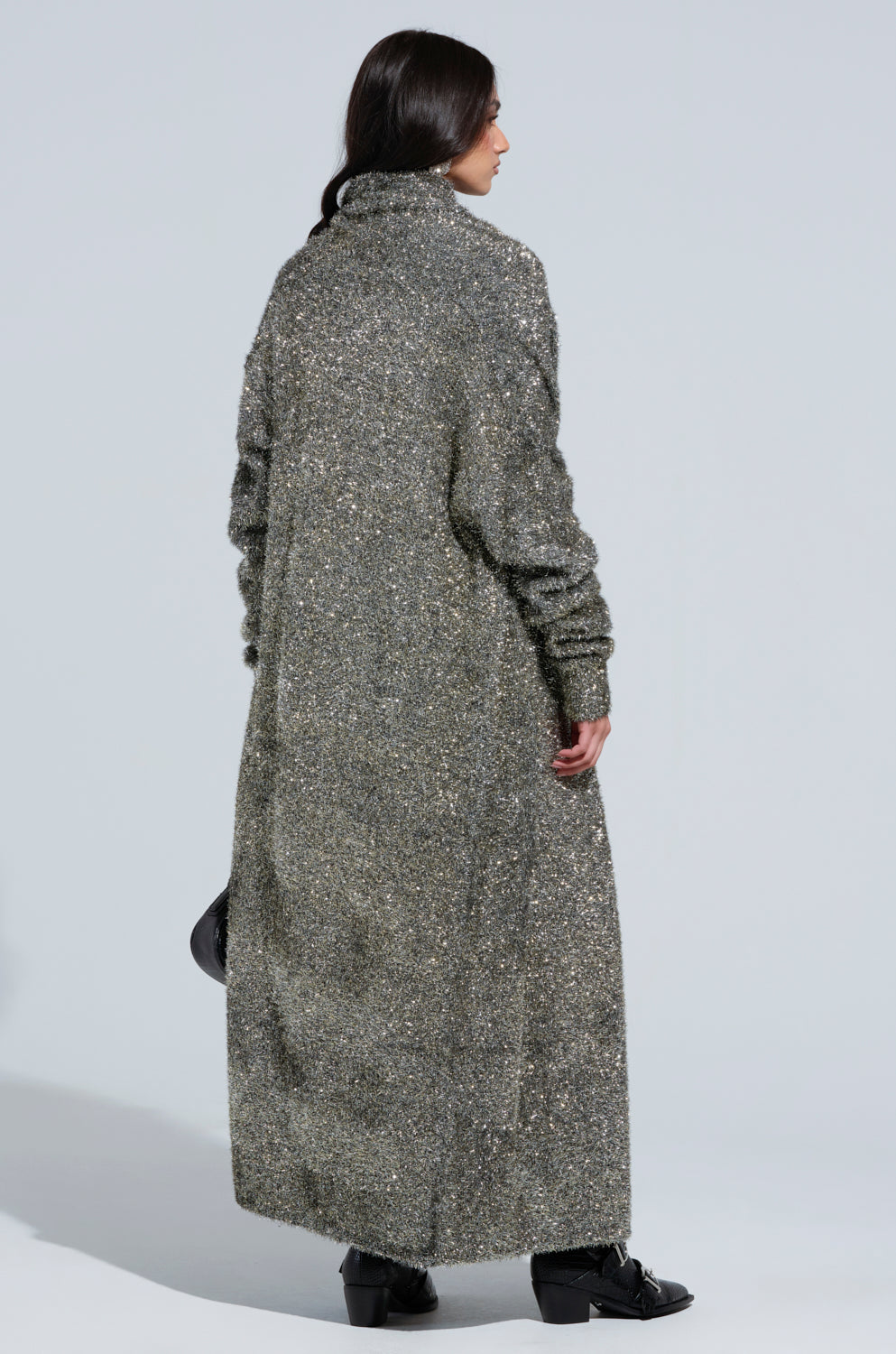 A person with long dark hair is wearing the WRAPPED IN GOLD LONG CARDIGAN, a textured gray piece from the AKIRA Label, featuring a high collar. They stand looking to the right, with the cardigan reaching their ankles and paired with black shoes. The plain and light background accentuates their striking style. 4