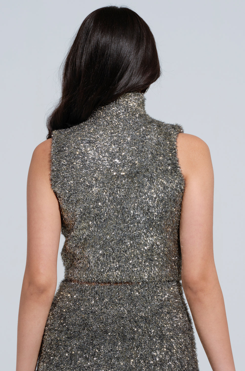 A person with long dark hair is seen from behind, wearing a sleeveless dress that features shimmering silver and gold tinsel fabric glistening in the light. The high collar adds elegance to the outfit, reminiscent of the style of the "WRAPPED IN GOLD CROPPED MOCK NECK SWEATER," against a plain, light-colored background. 2