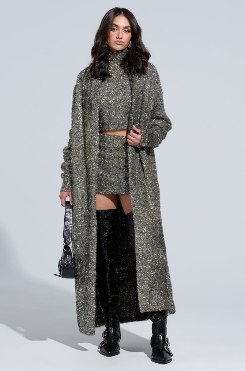 A woman dressed in a long, shimmering silver-textured gray overcoat paired with a matching crop top and skirt. She carries a small black bag and wears black knee-high boots featuring buckles. Her hair cascades in loose waves, styled elegantly. 4