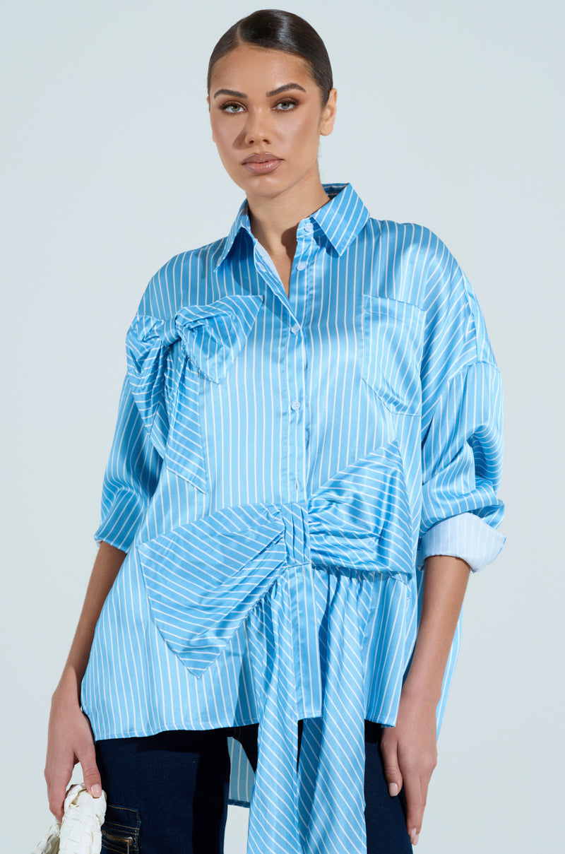 A woman in an oversized CLAIRE BIG BOW STRIPED BUTTON UP SHIRT, featuring light blue and white stripes with big bows, stands against a light background. Her hair is neatly tied back, and she holds a cream-colored item in her left hand.