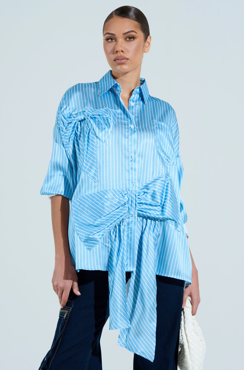 A person in a blue and white CLAIRE BIG BOW STRIPED BUTTON UP SHIRT, featuring prominent bow details, gathered fabric, and an oversized fit, poses against a plain background. They accessorize with a white clutch and dark pants.