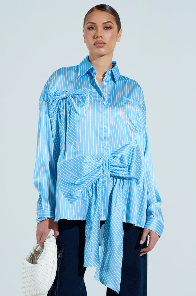 Wearing the oversized CLAIRE BIG BOW STRIPED BUTTON UP SHIRT, paired with dark pants and a textured white handbag, a person sports a sleek bun against a solid light background.