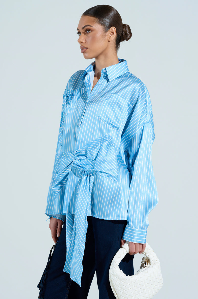 Sporting the CLAIRE BIG BOW STRIPED BUTTON UP SHIRT in a shiny, oversized fit, a person stands against a light background. They carry two bags, one white and one black, with their hair styled in a sleek bun.