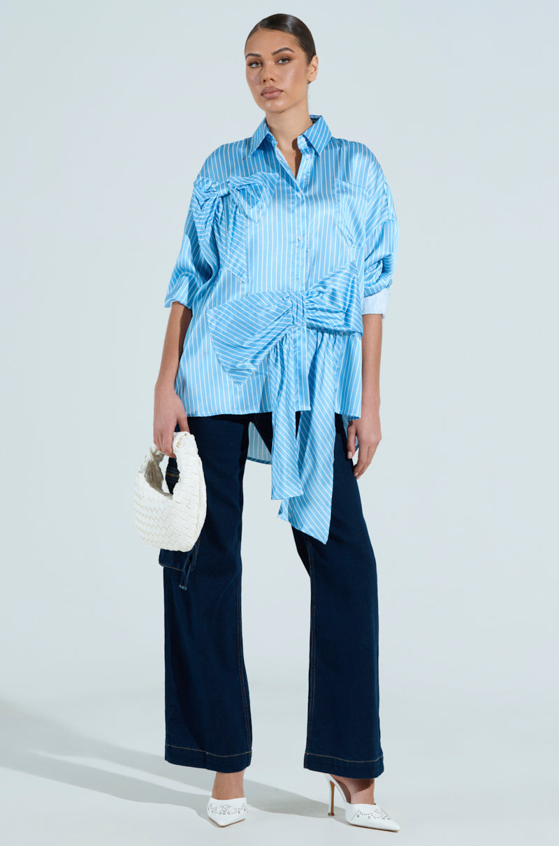 A person wears the CLAIRE BIG BOW STRIPED BUTTON UP SHIRT with dark wide-leg jeans, carries a white woven handbag, and sports white pointed-toe heels, all complemented by a sleek bun hairstyle.