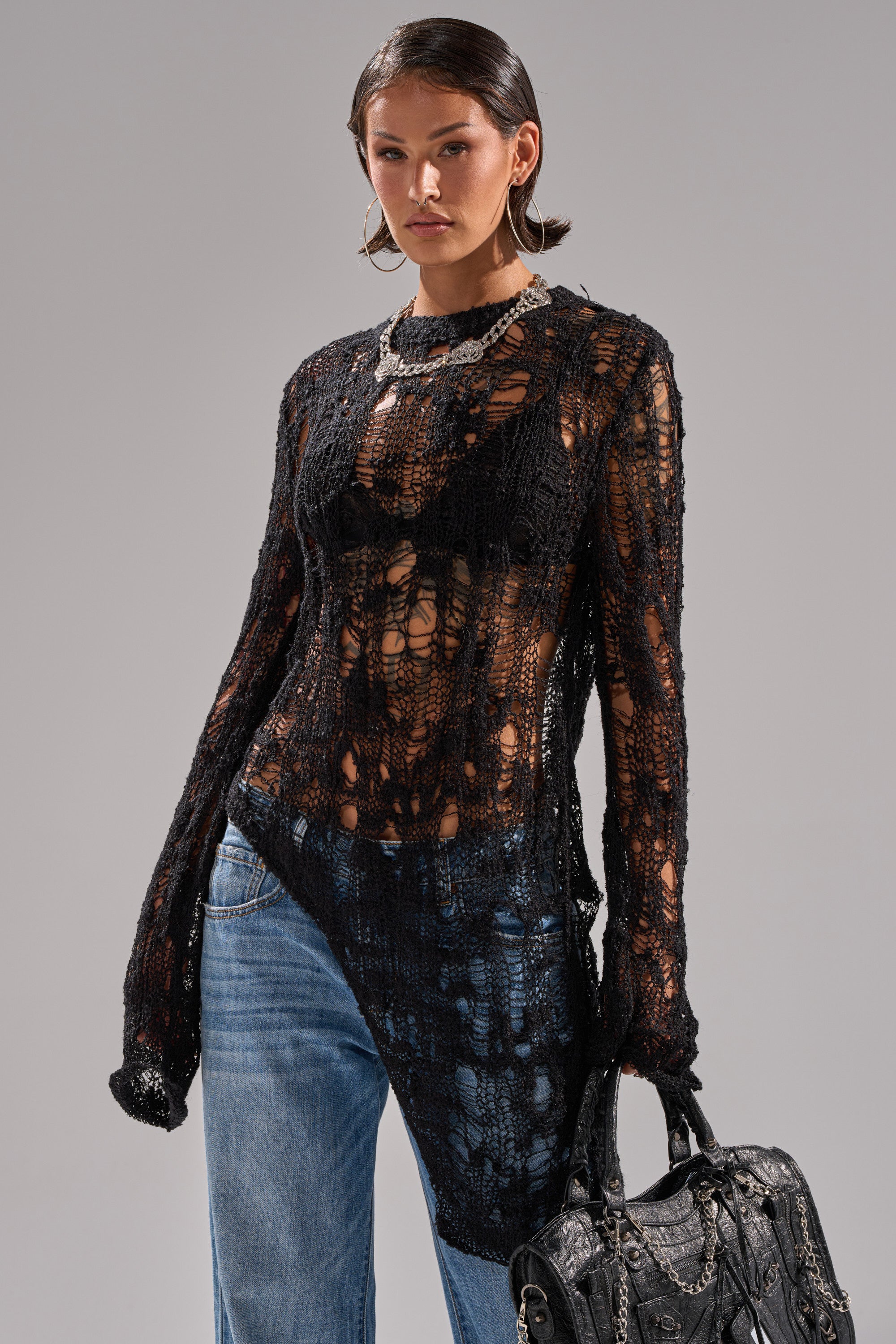 A woman poses in the ASYMMETRICAL KNIT SWEATER, a black sheer top, styled over a black bra with light blue jeans, hoop earrings, a chain necklace, and a textured black handbag against a plain background.