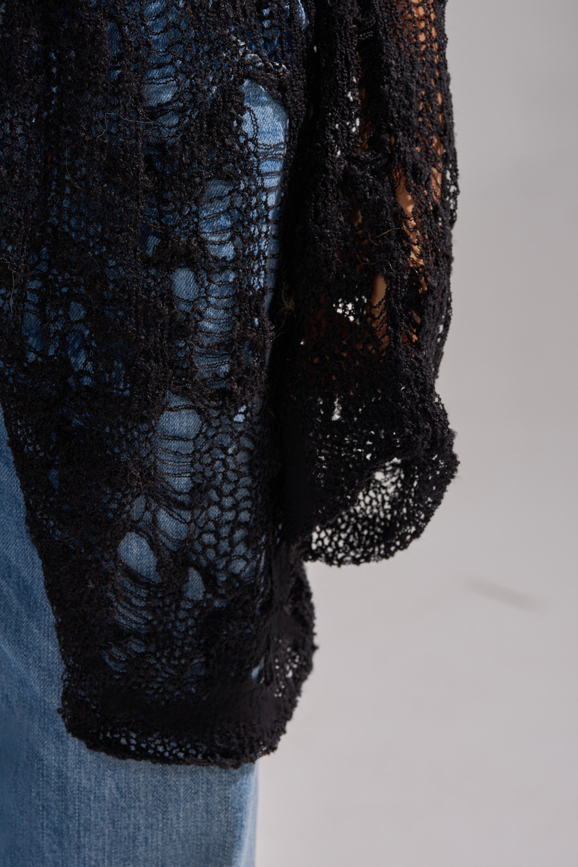 Close-up of a person wearing blue jeans and the ASYMMETRICAL KNIT SWEATER, a black distressed style draped over their leg, set against a neutral gray background.