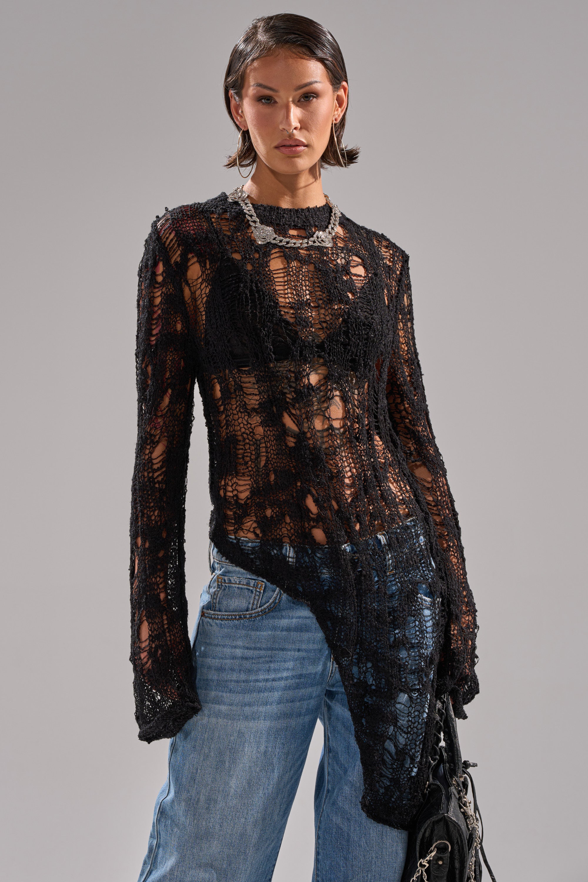 A woman models the ASYMMETRICAL KNIT SWEATER in black over a bralette and blue jeans, styled with a chunky silver necklace and hoop earrings, against a gray background.