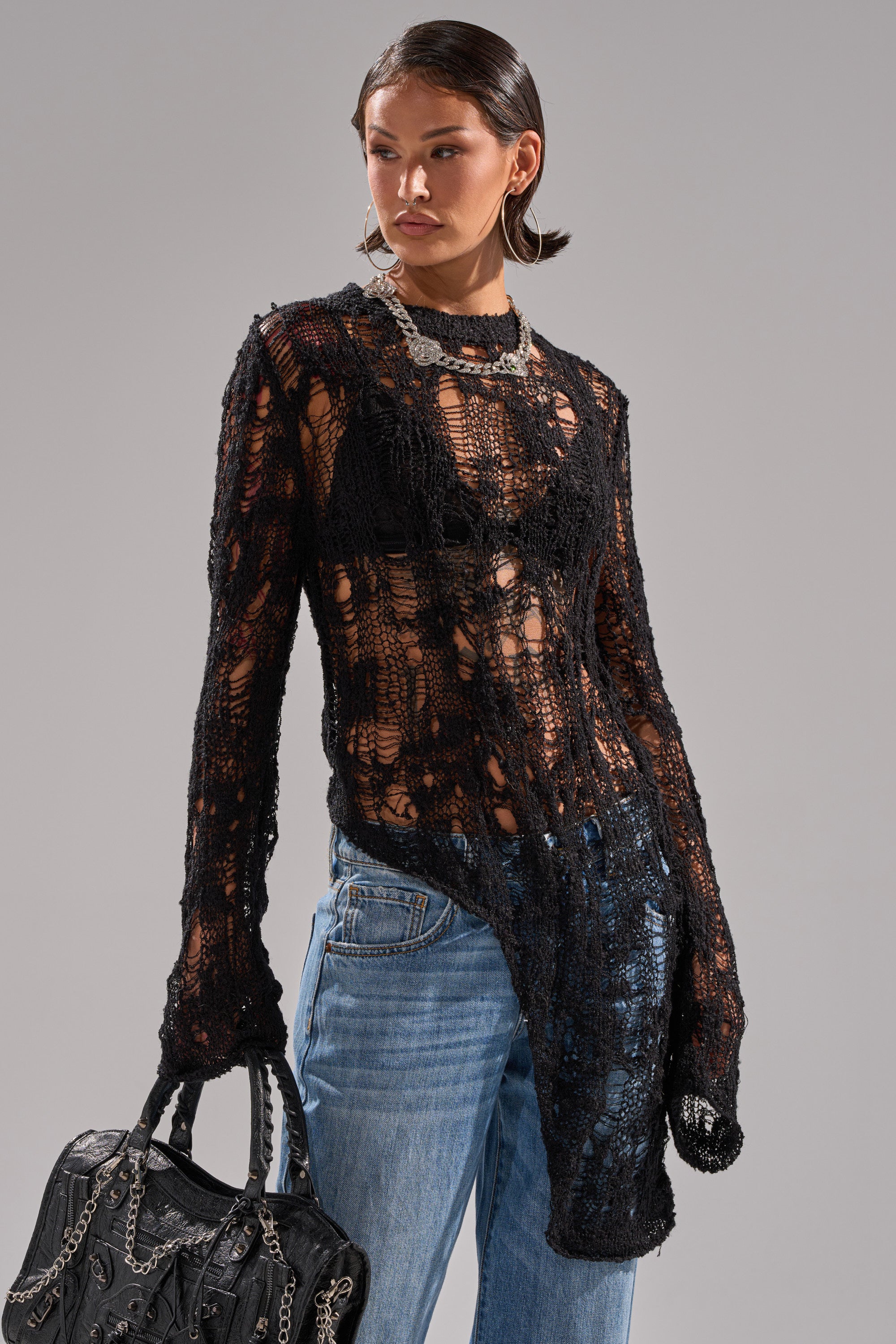 A woman models the ASYMMETRICAL KNIT SWEATER in sheer black over a black bra with blue jeans. She accessorizes with a black chain-detailed bag and chunky jewelry, posing against a plain gray backdrop.