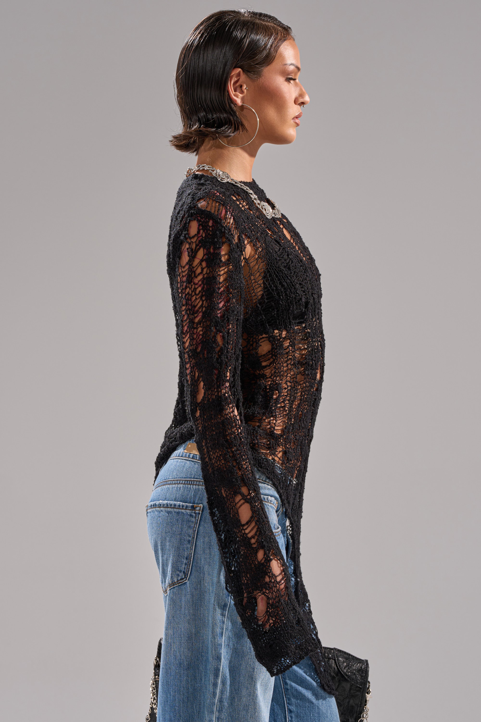 A person with slicked-back hair wears the ASYMMETRICAL KNIT SWEATER—a sheer, black, distressed long-sleeve style—paired with light blue jeans. They are shown in profile against a plain gray background.