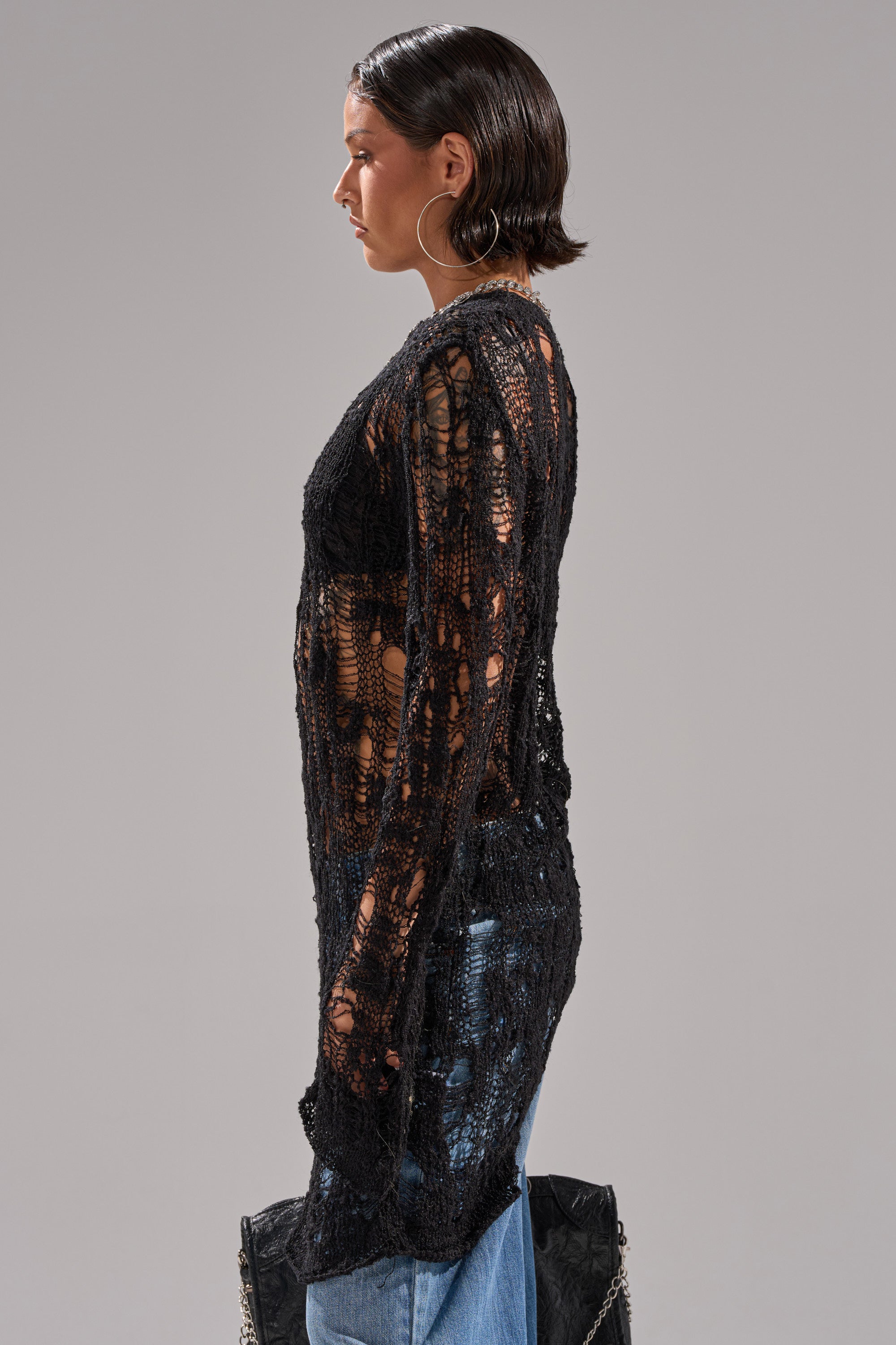 A person with short, slicked-back dark hair stands in profile against a light gray background, wearing the ASYMMETRICAL KNIT SWEATER in black over blue jeans, large hoop earrings, and holding a black bag by their side.