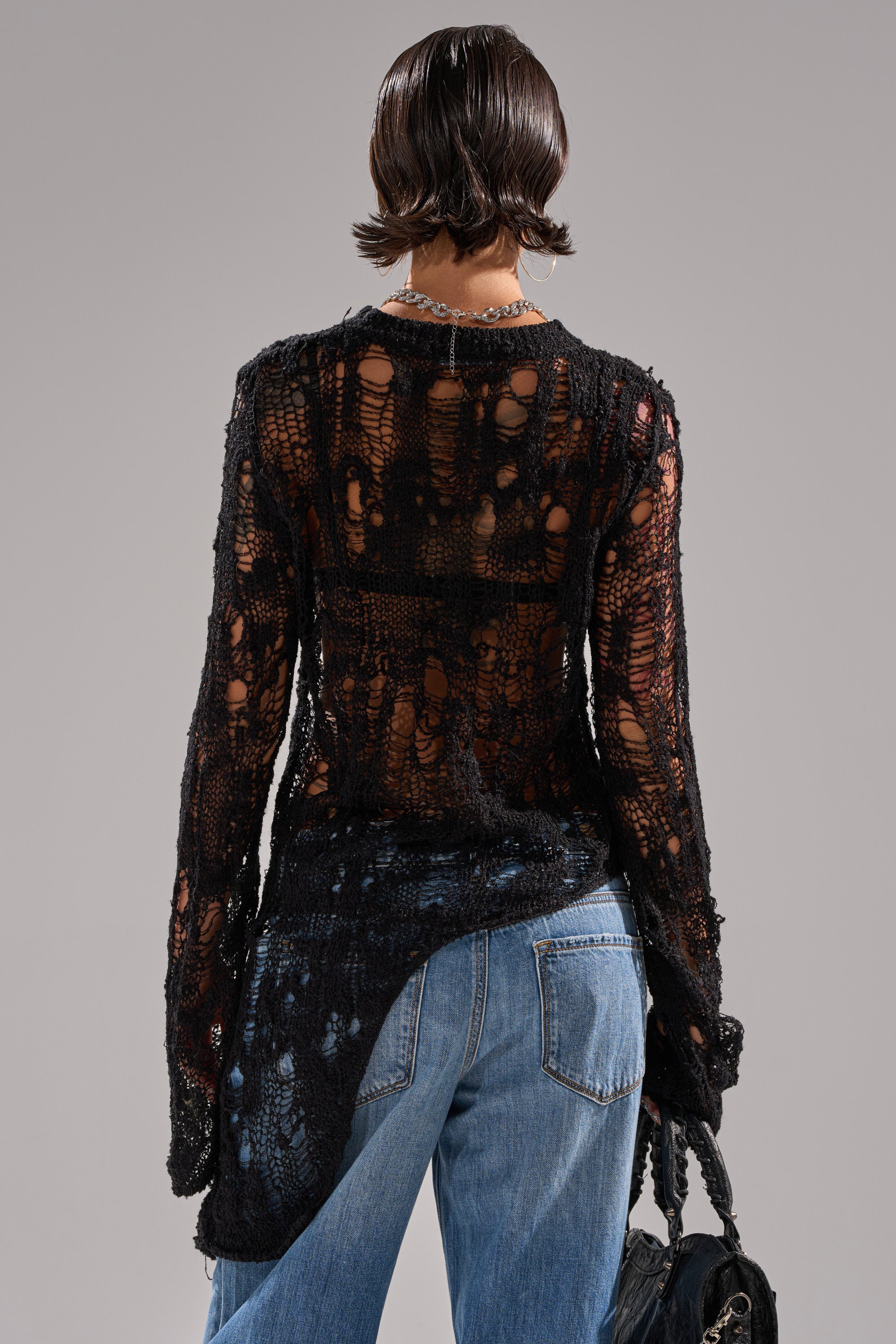 A person stands facing away in the ASYMMETRICAL KNIT SWEATER, a black, distressed knit with an asymmetrical design, styled over blue jeans. They have short, slicked-back hair and hold a black bag in their left hand against a plain gray background.