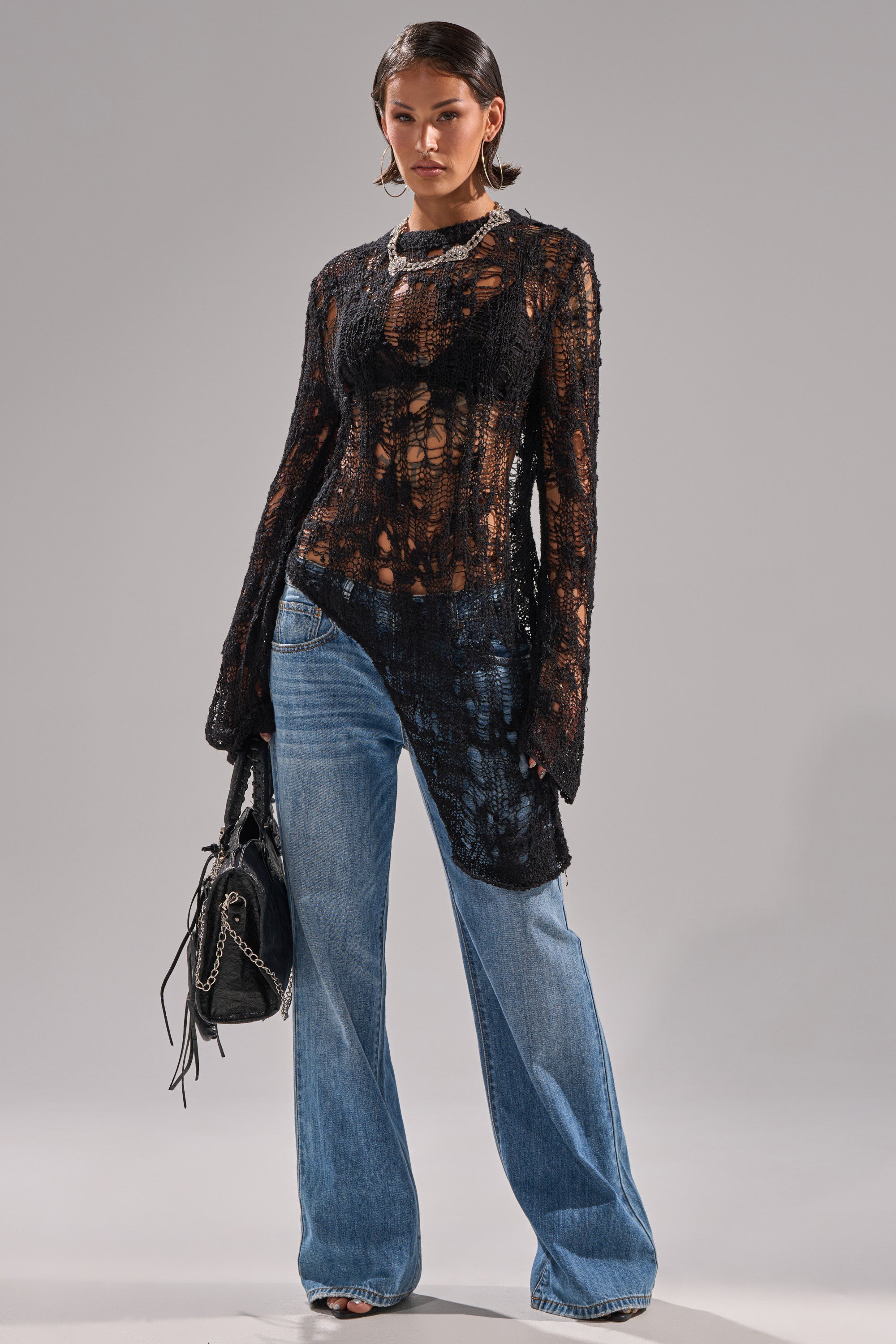 A woman models the ASYMMETRICAL KNIT SWEATER in black with distressed details and an uneven hem, paired with wide-leg blue jeans, silver heels, hoop earrings, and a black handbag against a plain gray background.