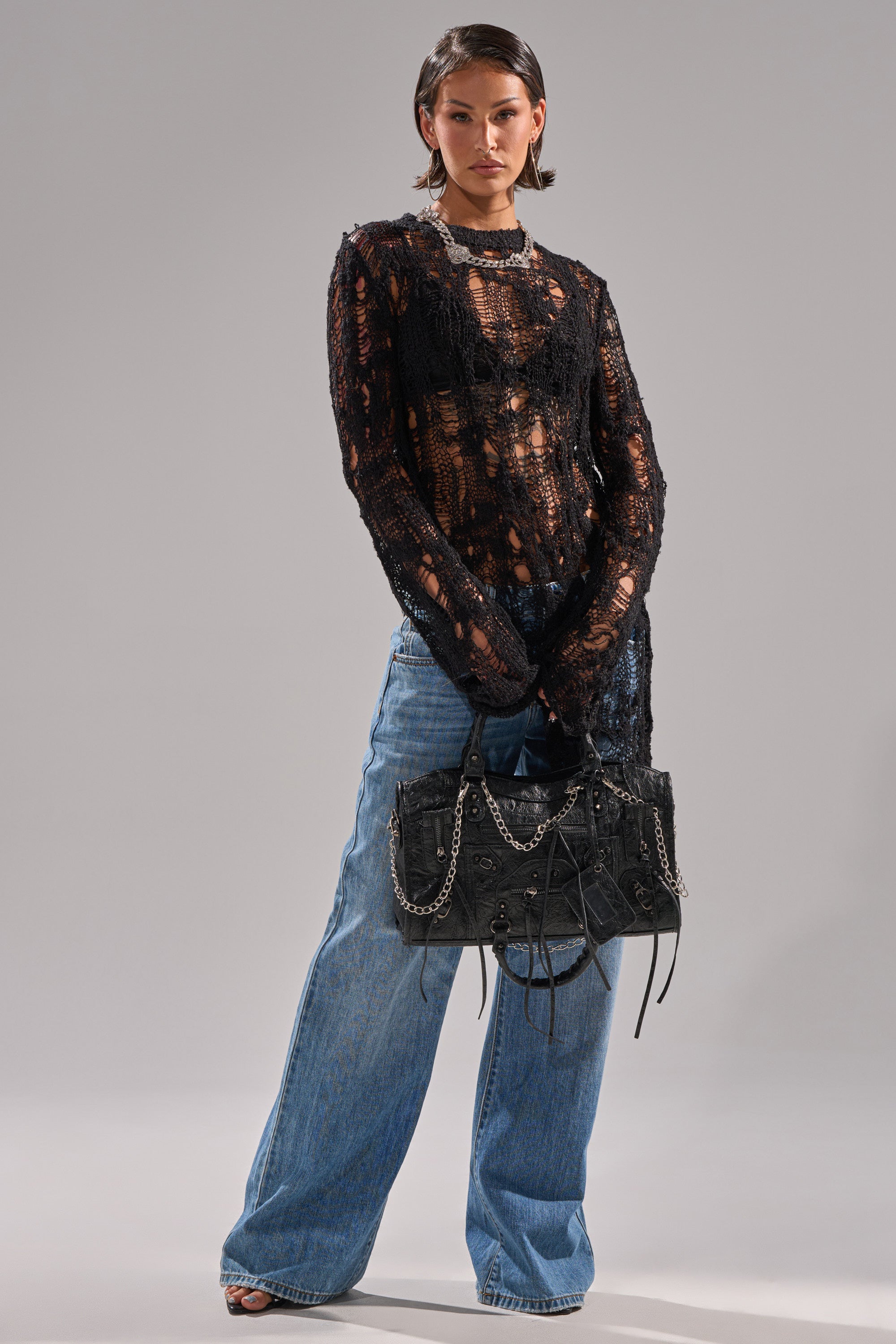 A person with short, slicked-back hair and a neutral expression stands against a gray background, wearing an ASYMMETRICAL KNIT SWEATER, loose blue jeans, and holding a textured black handbag with chain straps.