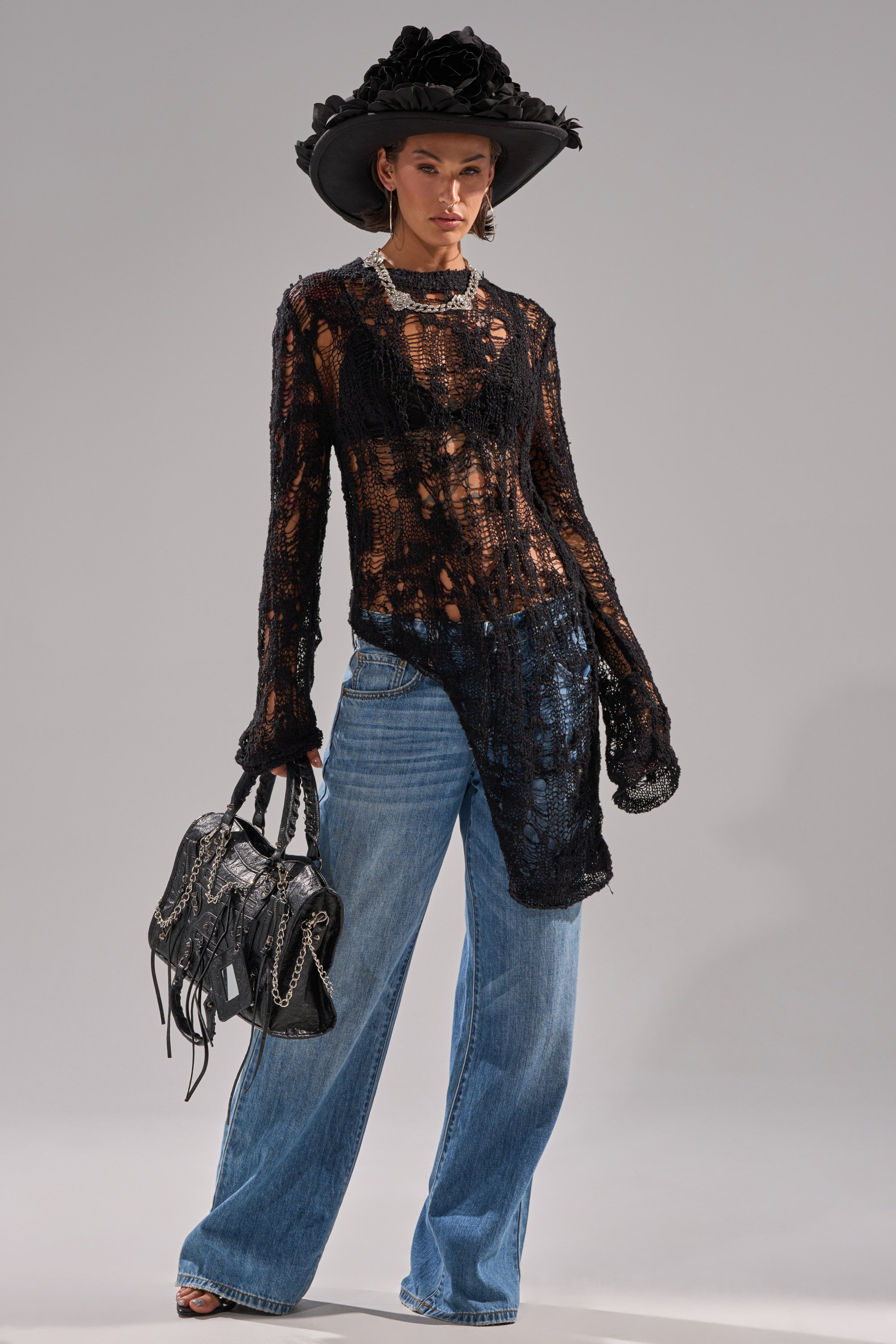 Someone styled the ASYMMETRICAL KNIT SWEATER over a black bra, pairing it with wide-leg blue jeans, a large black floral hat, silver jewelry, and a black fringed handbag.