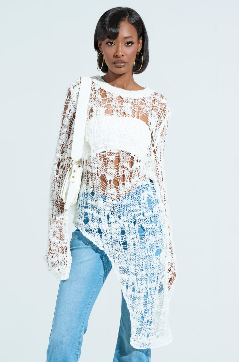 Wearing an asymmetrical knit top layered over a tank, paired with light blue jeans, the person completes their look with large hoop earrings and a white shoulder bag. The gauzy material of the top stands out against the plain, bright background.