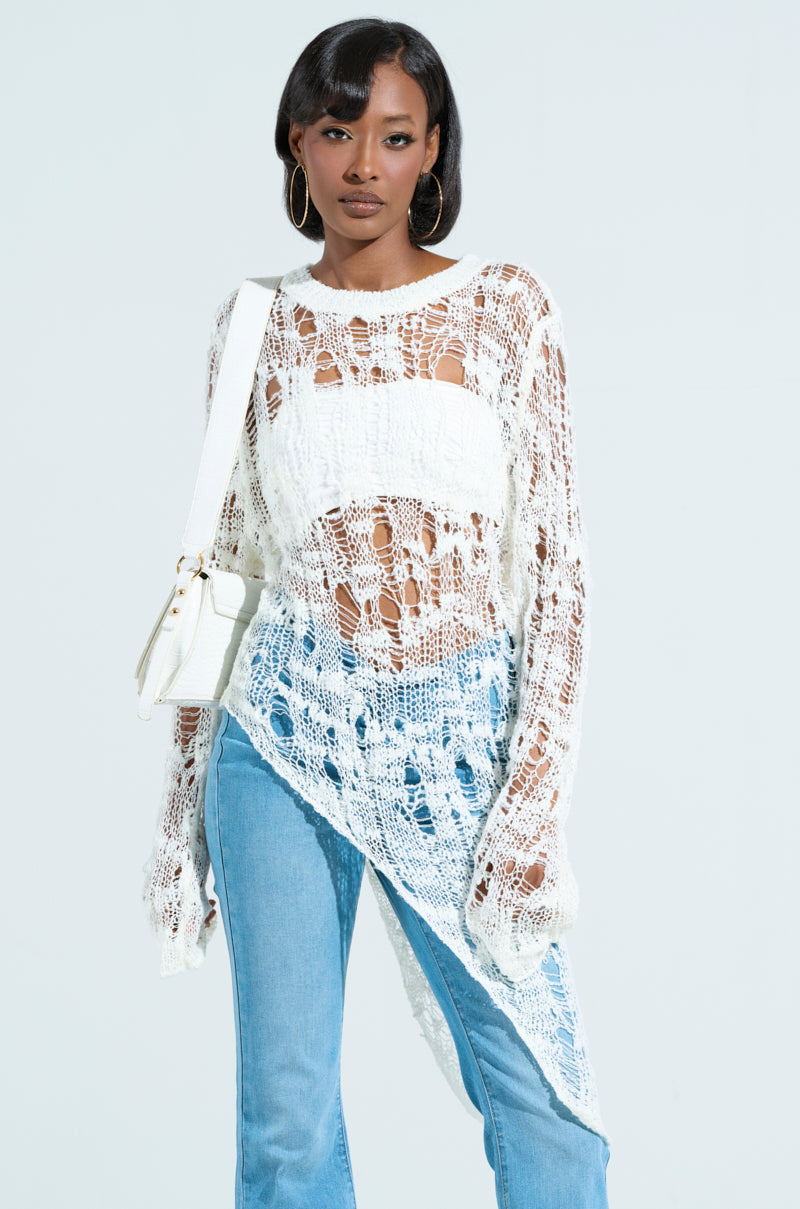 Someone wearing an ASYMMETRICAL KNIT TOP over a white tank matches it with light blue jeans. They hold a white shoulder bag and wear hoop earrings, standing against a light background.