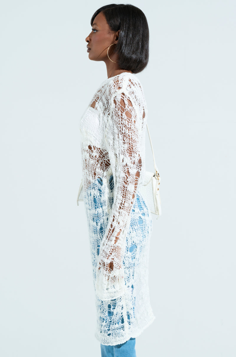 Side profile of a person wearing an ASYMMETRICAL KNIT TOP, complemented by a long crochet cardigan over white and blue jeans. They sport short black hair, large hoop earrings, and carry a small white bag against a plain background.