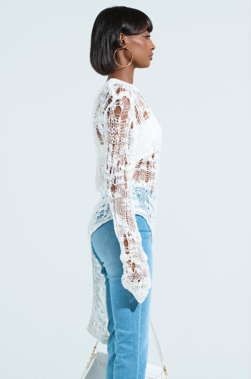 A woman in side profile wears an ASYMMETRICAL KNIT TOP with a tank and light blue jeans. She accessorizes with large hoop earrings and carries a white handbag against a plain background.
