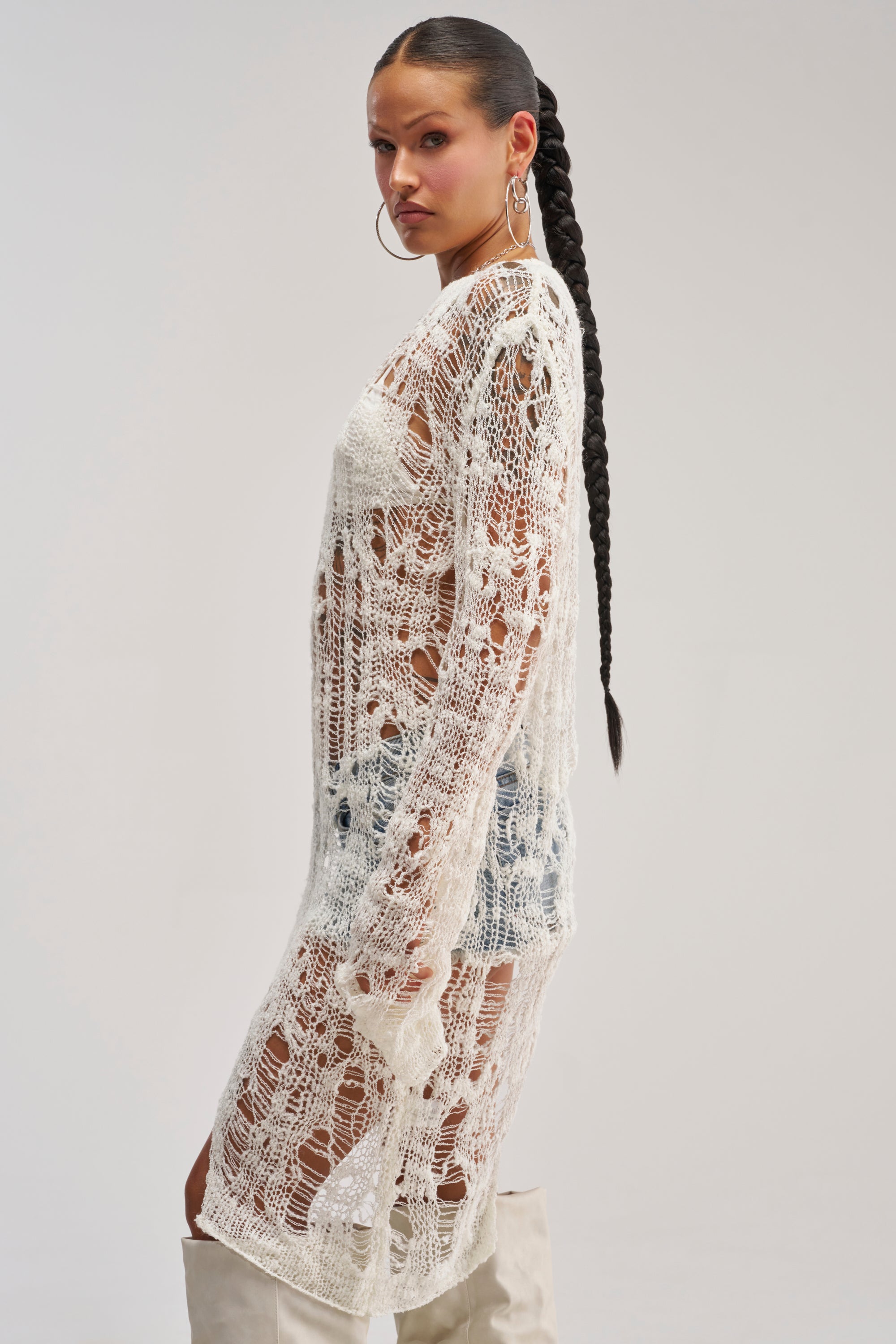A woman with long braided hair and large hoop earrings showcases the ASYMMETRICAL KNIT TOP IN IVORY—a sheer, white, asymmetrical piece styled over denim shorts and a top. She stands sideways against a plain light background, bringing a fresh twist to streetwear with subtle ripped detailing for an edgy effect.