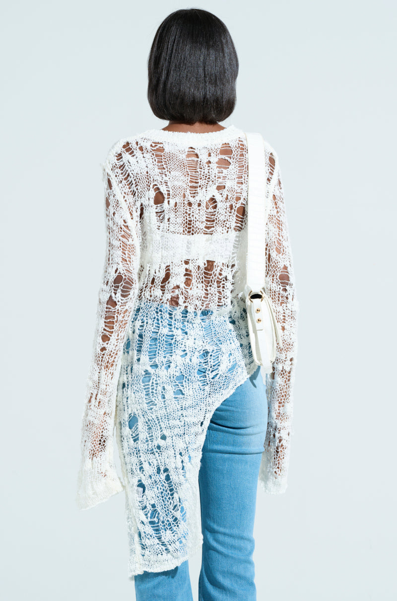 Wearing the ASYMMETRICAL KNIT TOP with frayed edges over blue jeans, a person with shoulder-length hair faces away, carrying a white shoulder bag on their right. The background is a plain light color.