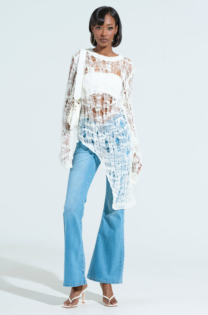 A person wearing an ASYMMETRICAL KNIT TOP in sheer white with a web-like texture, light blue flared jeans, white high-heeled sandals, and a white shoulder bag stands against a plain background.