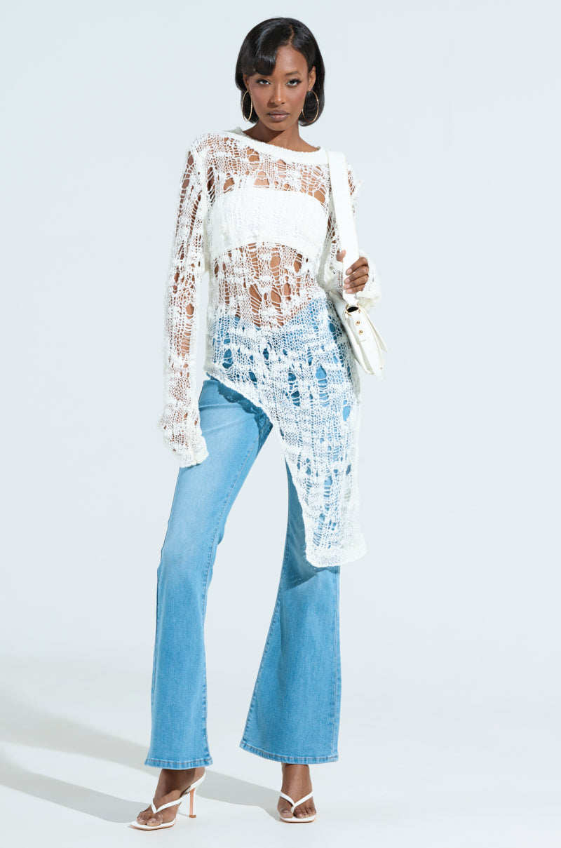 A person in an ASYMMETRICAL KNIT TOP over a white bandeau pairs it with light blue flared jeans, complemented by white high heels and a matching shoulder bag. The backdrop is plain white.