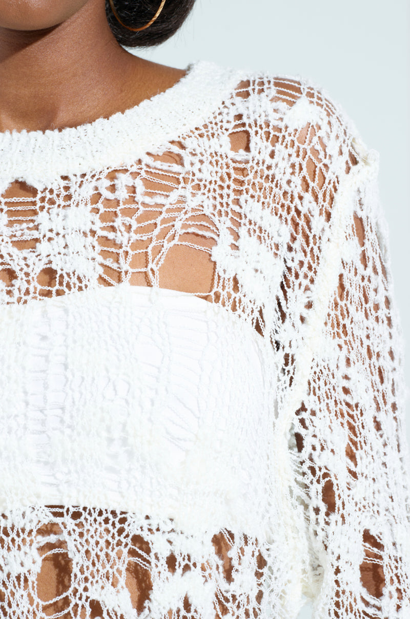 Close-up of a person wearing an ASYMMETRICAL KNIT TOP over a white bandeau, showcasing its delicate, web-like crochet pattern with both open and tight weaves. The image highlights the shoulder and upper torso area.
