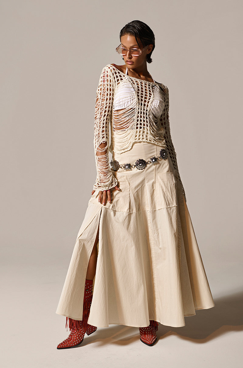 A person stands confidently in a GINA LONG KNIT TOP with a white scoop neckline over a chic outfit, accessorized by a floral-designed wide belt. Red fringe-detailed ankle boots complete the look against a neutral background.