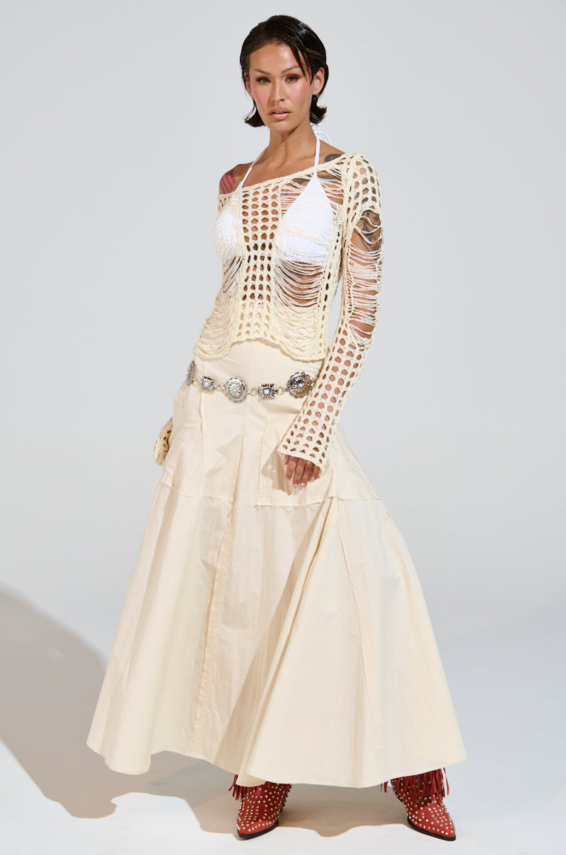 The ensemble features the GINA LONG KNIT TOP, a cream crochet knit with scoop neckline, elegantly worn over a white bikini and paired with a long cream skirt and decorative belt. Red patterned boots complete the look, all set against a plain white backdrop.