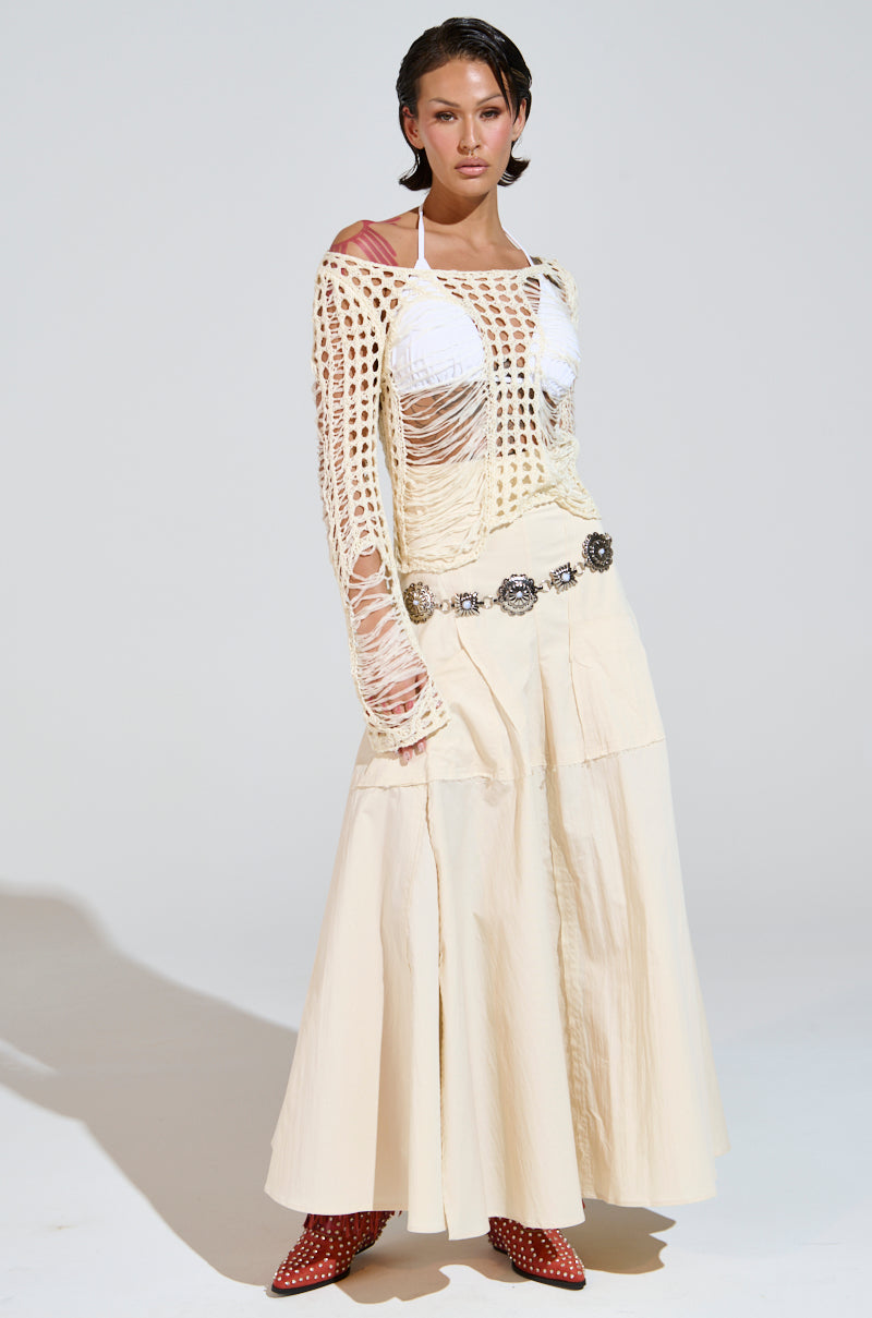A person poses in a cream-colored, long skirt paired with the GINA LONG KNIT TOP featuring a scoop neckline and loose, net-like overlay. They accessorize with a silver belt and red boots against a plain, light-toned background.
