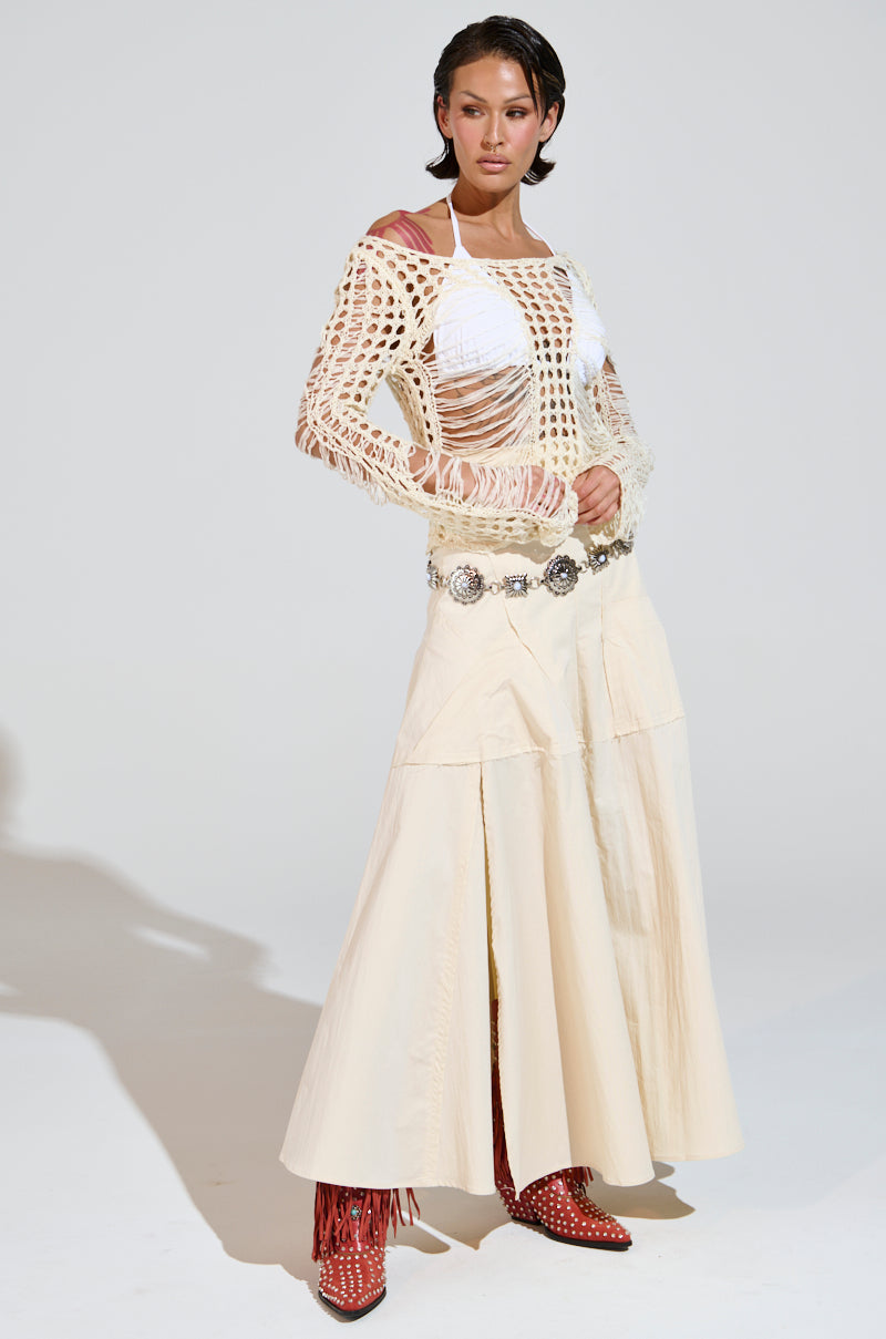 A woman confidently poses in the GINA LONG KNIT TOP with distressed detailing over a white shirt, paired with a long cream skirt. Her look is accessorized by a silver belt and bold red studded ankle boots. Her hair is styled back as she stands against a plain backdrop.