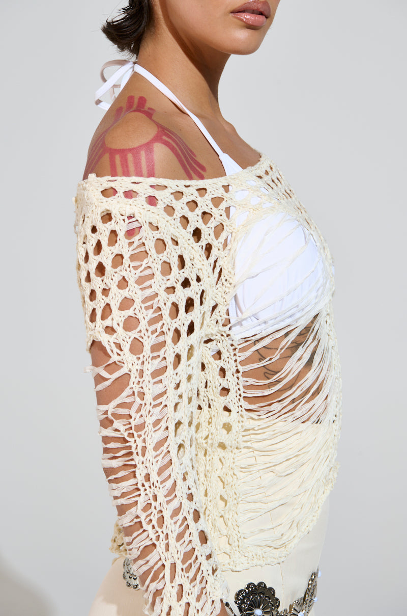 Wearing the white GINA LONG KNIT TOP, a person stands sideways, the crochet cover-up revealing their bikini beneath. The scoop neckline frames their figure while a pink geometric tattoo decorates their shoulder against a plain light gray background.