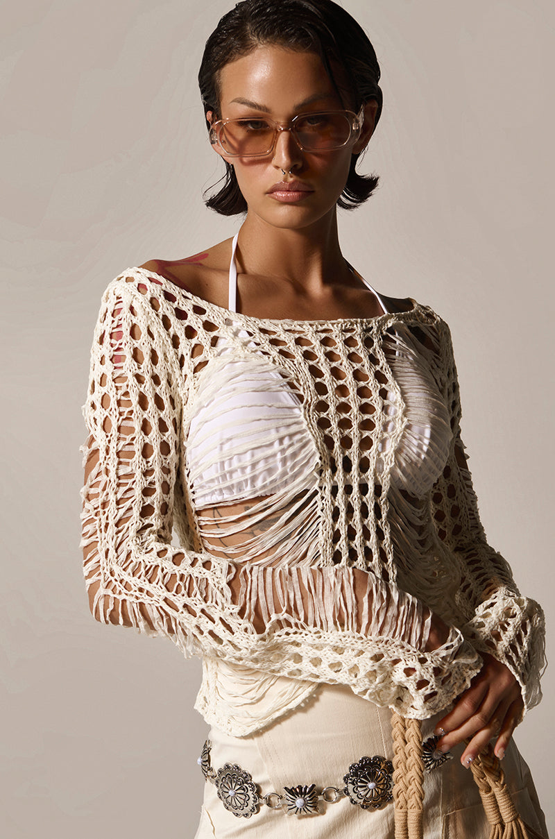 The focal point is a person in a GINA LONG KNIT TOP with distressed detailing over a white bikini. They have short, dark hair and stylish sunglasses, complemented by a patterned belt on a light skirt. The plain backdrop enhances their chic summer look.
