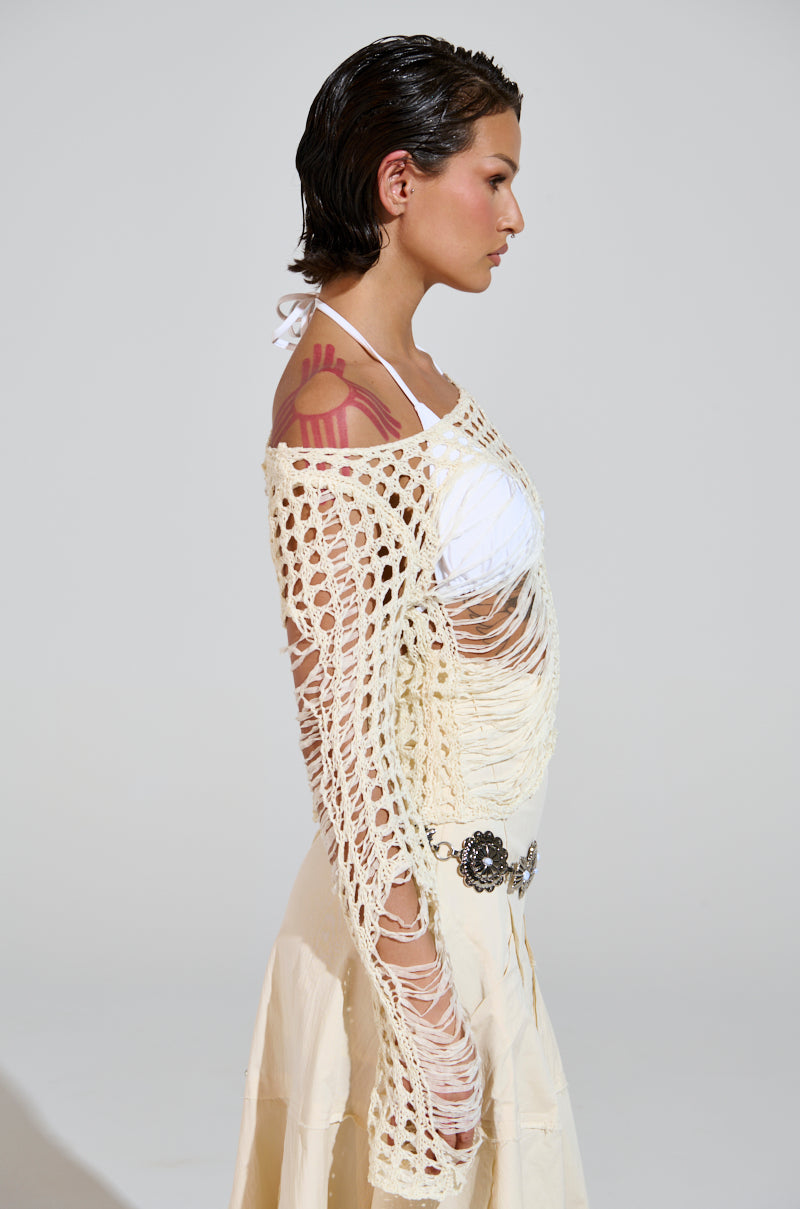 In profile, the person sports slicked-back hair in a white halter beneath the GINA LONG KNIT TOP, a woven beige mesh sweater. Their white skirt with a decorative belt and shoulder tattoo stands out against the plain background, emphasizing the chic ensemble's distressed detailing.