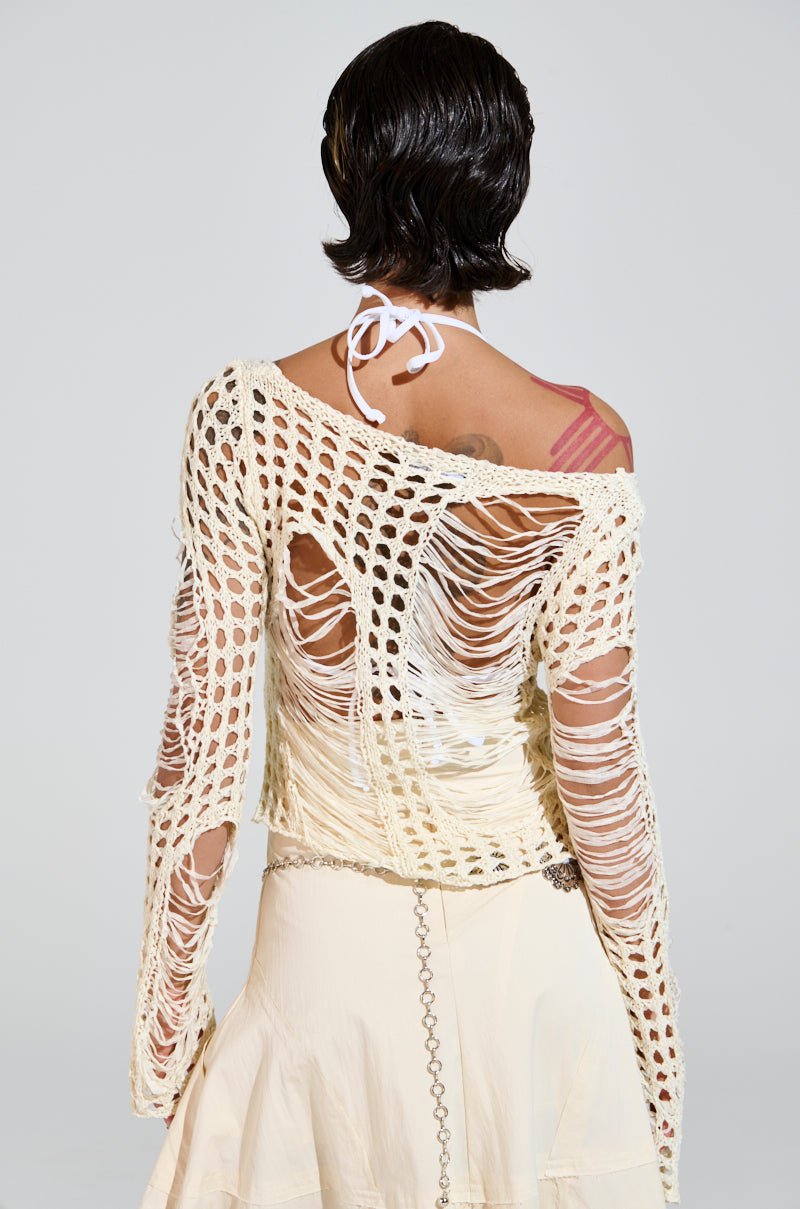 A person with short black hair, seen from behind, wears the GINA LONG KNIT TOP over a scoop neckline halter. Their white skirt complements a chain belt elegantly, while their tattooed shoulder adds flair. The light-colored backdrop highlights the unique texture of their outfit.
