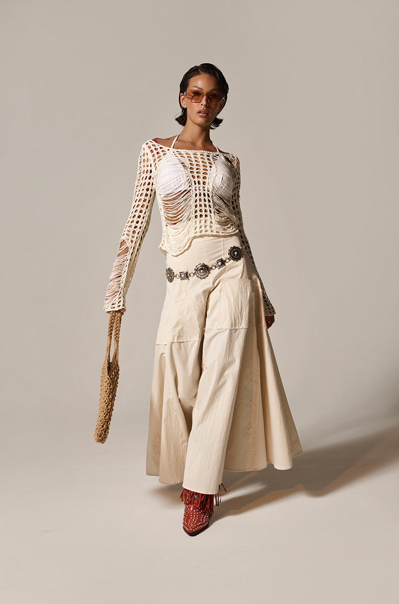 A person confidently poses in the GINA LONG KNIT TOP with distressed details over a white bralette, paired with beige wide-leg pants, red shoes, sunglasses, a large silver belt, and holding a woven tote bag against a plain background.