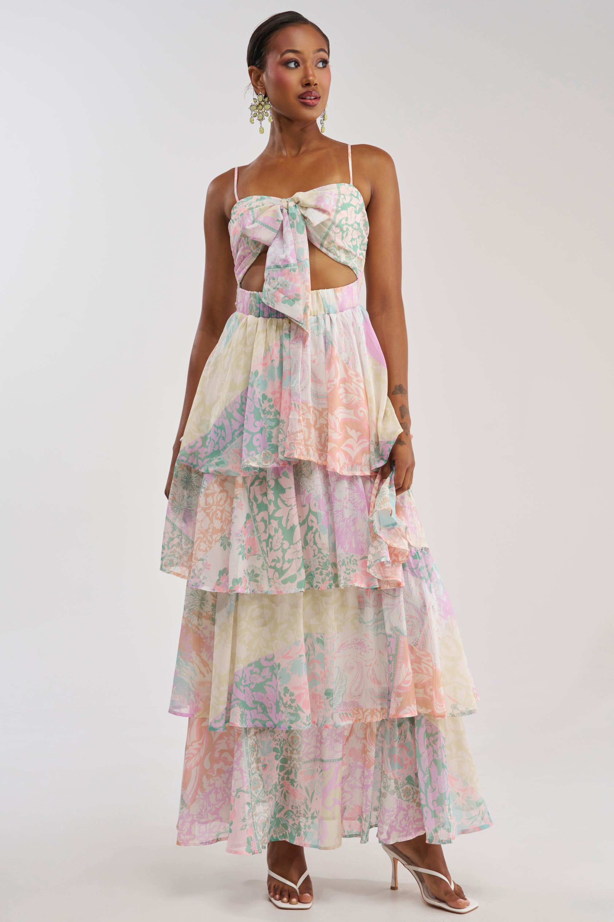 A woman wearing the DAY DREAMER MULTICOLORED CHIFFON MAXI DRESS, featuring a pastel floral print, large front bow and cutout detail, spaghetti straps, and paired with strappy white heels, poses against a plain white background—a beautiful choice for a summer wedding dress.