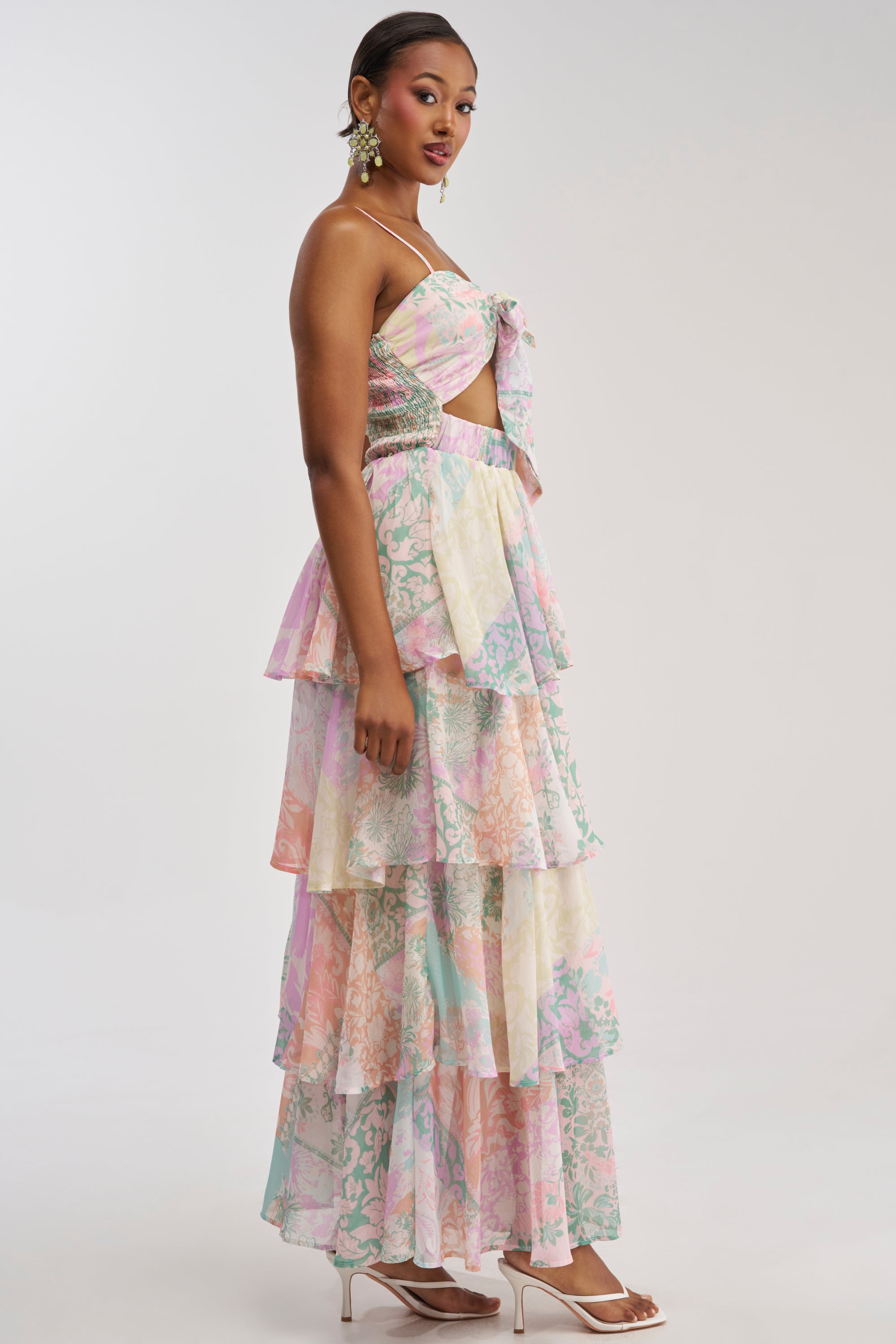 A woman wearing the DAY DREAMER MULTICOLORED CHIFFON MAXI DRESS, featuring pastel florals, side cutouts, and a bow detail, paired with white heeled sandals and gold earrings, stands against a plain light background—making it the perfect choice for a summer wedding.