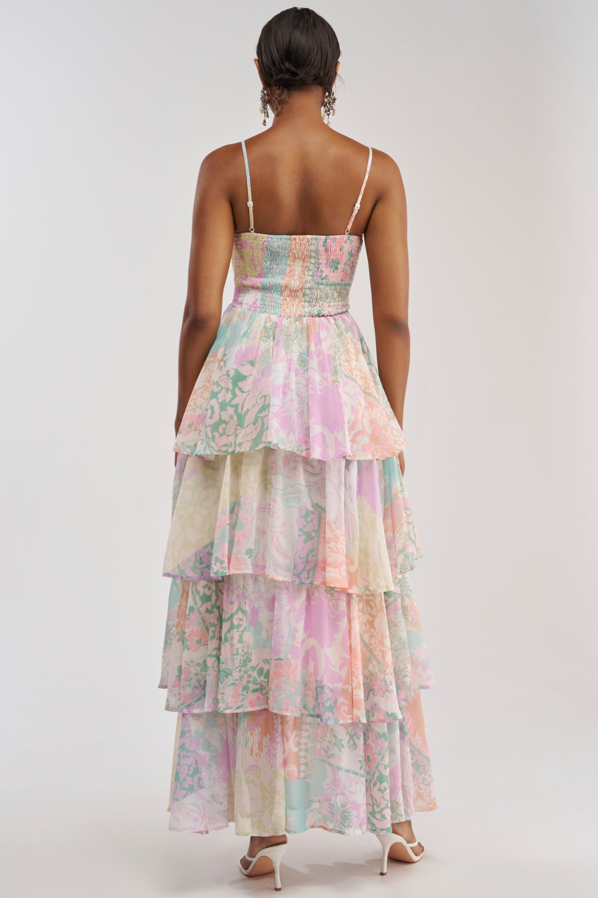A woman faces away, wearing the DAY DREAMER MULTICOLORED CHIFFON MAXI DRESS—featuring thin straps and tiered ruffled layers in pastel floral chiffon—paired with white heels, against a plain light background.