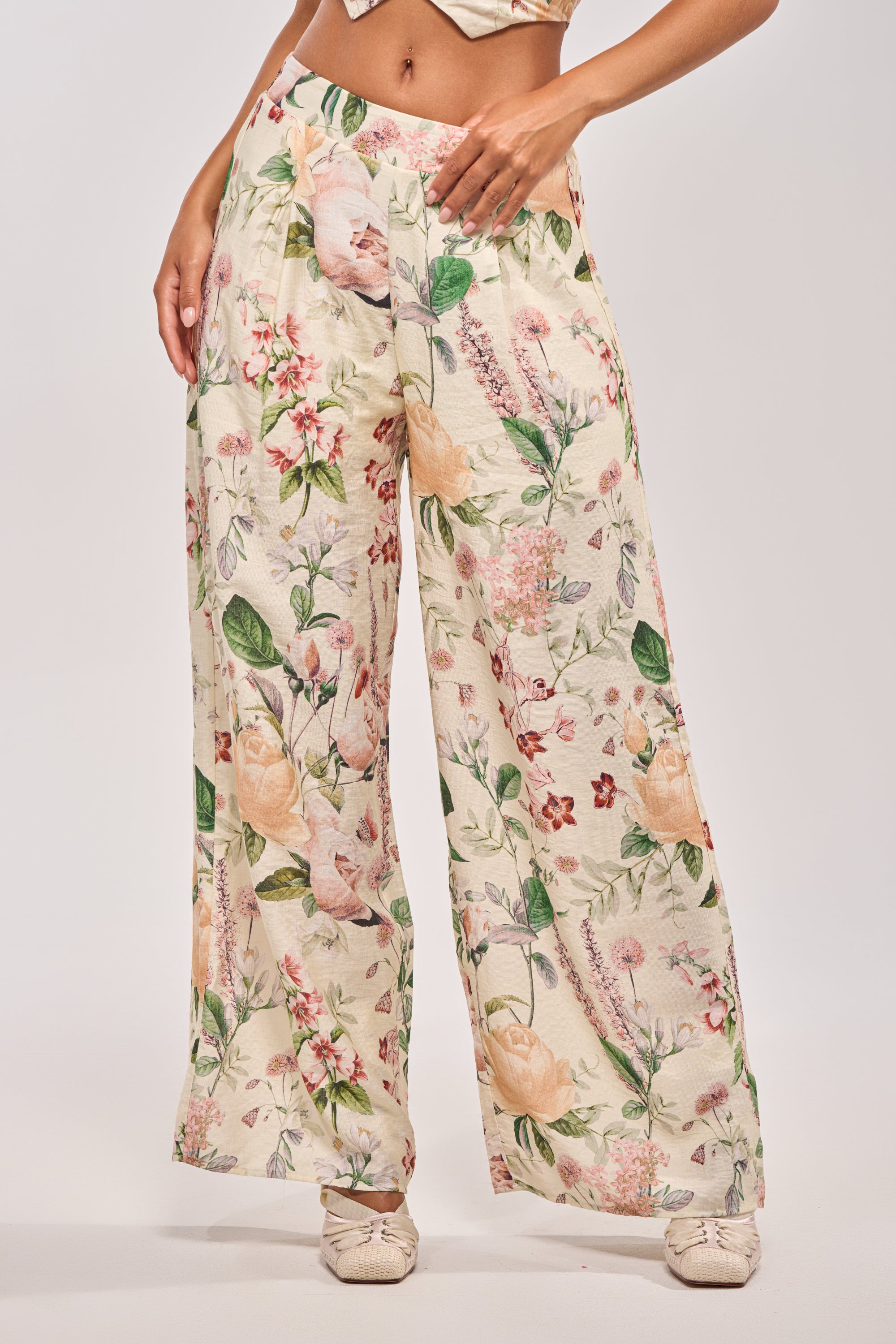 Wearing the LADY B TROUSER—loose, wide-leg, high-rise pants with a floral print in cream, pink, green, and yellow—a person pairs them with light shoes and stands against a plain background.