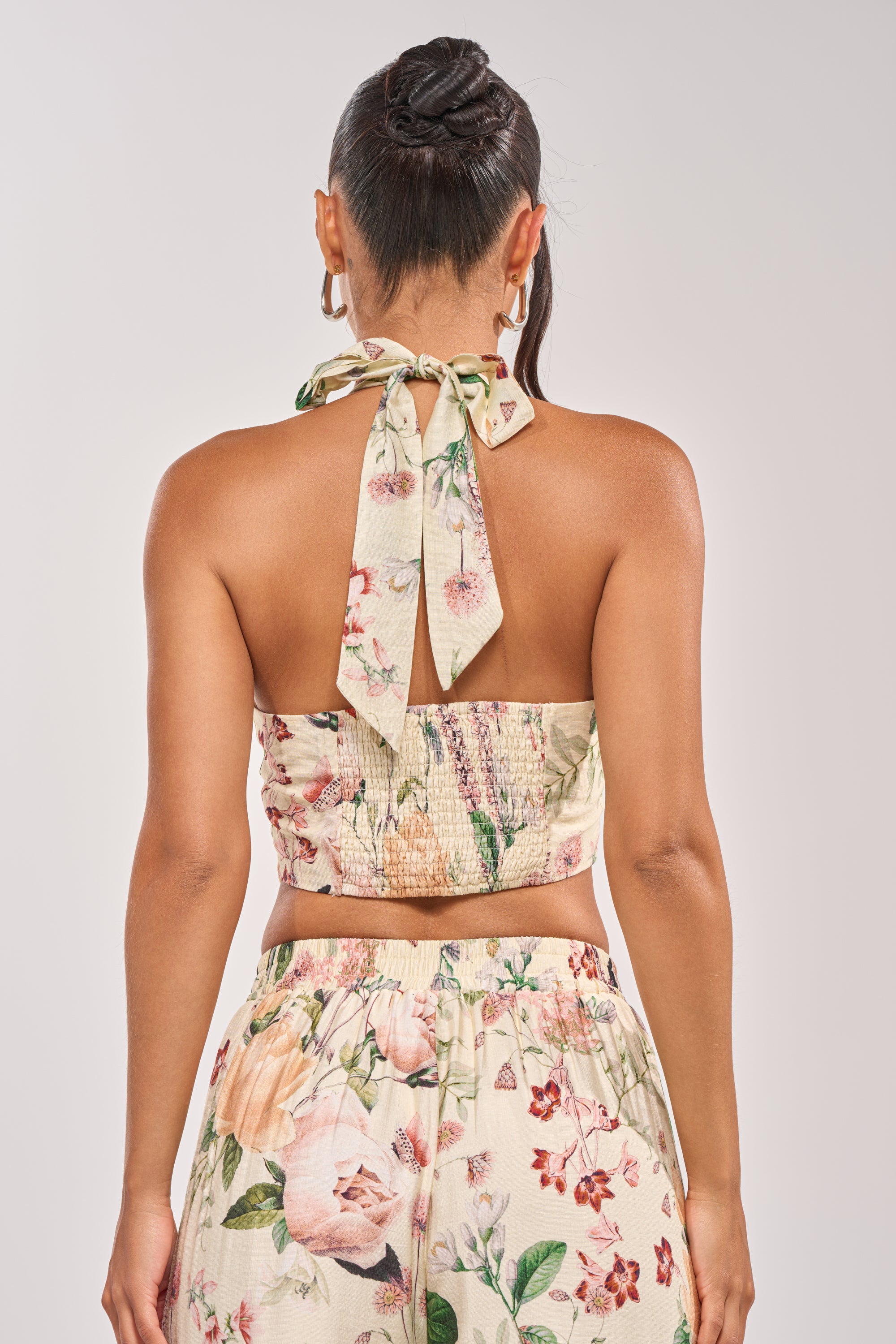 A woman is seen from behind wearing the LADY B BUTTON DOWN TANK, a halter, button-down top tied at the neck with matching high-waisted pants. Both pieces have a cream background and pink and green floral print. 4