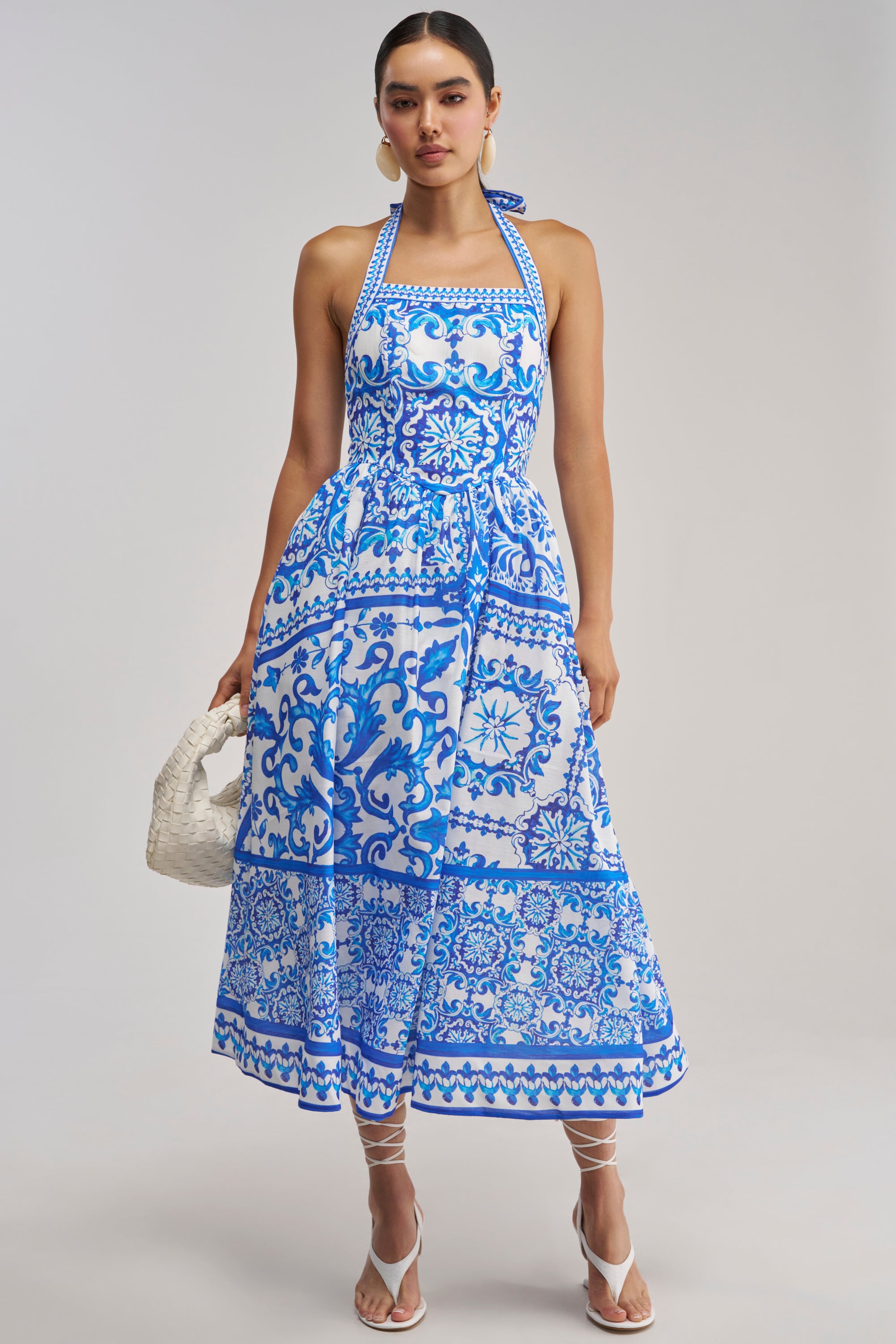 A model poses in the COCO CRUSH PRINTED HALTER MIDI DRESS, showcasing its blue and white Mediterranean-inspired floral tile pattern. The dress features a halter neckline and a flattering midi length—perfect for summer. She completes her look with white strappy sandals and a white woven handbag, set against a plain grey background.