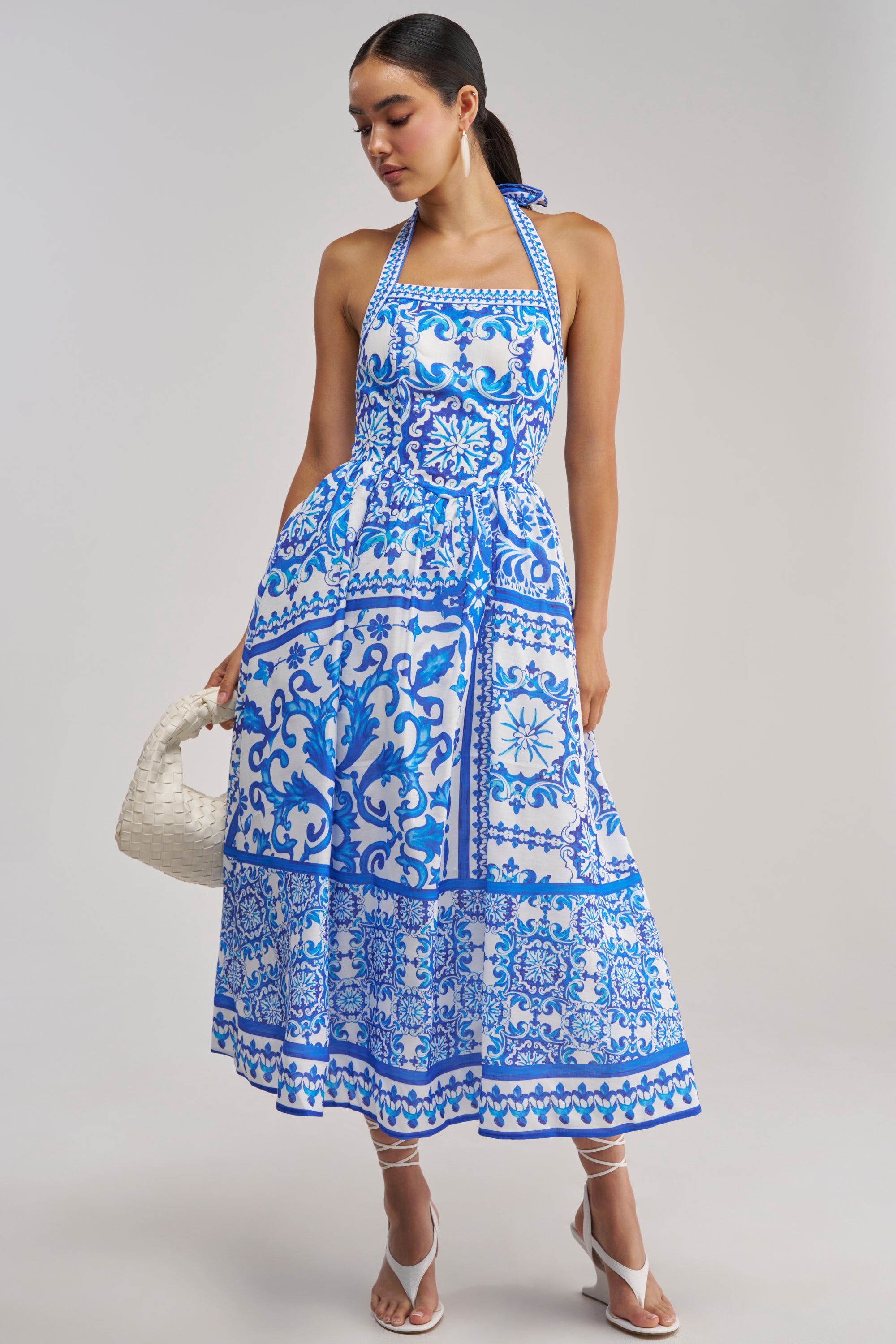 A woman wears the COCO CRUSH PRINTED HALTER MIDI DRESS—a blue and white floral tile print halter midi dress—while holding a white woven bag and standing in white heeled sandals against a plain background.