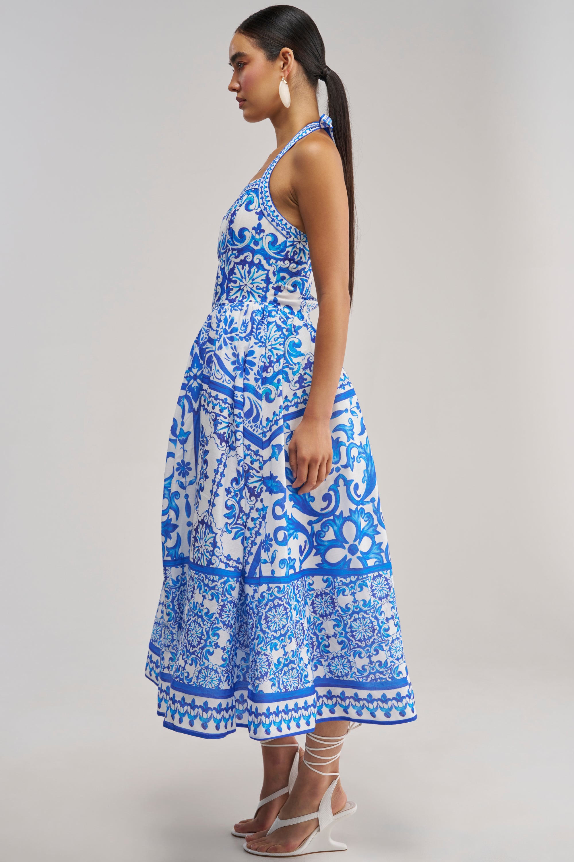 A woman stands sideways against a plain light background, wearing the COCO CRUSH PRINTED HALTER MIDI DRESS—a blue and white floral tile-print midi dress with a halter neck and ankle-length silhouette—paired with white strappy heels and large white earrings.