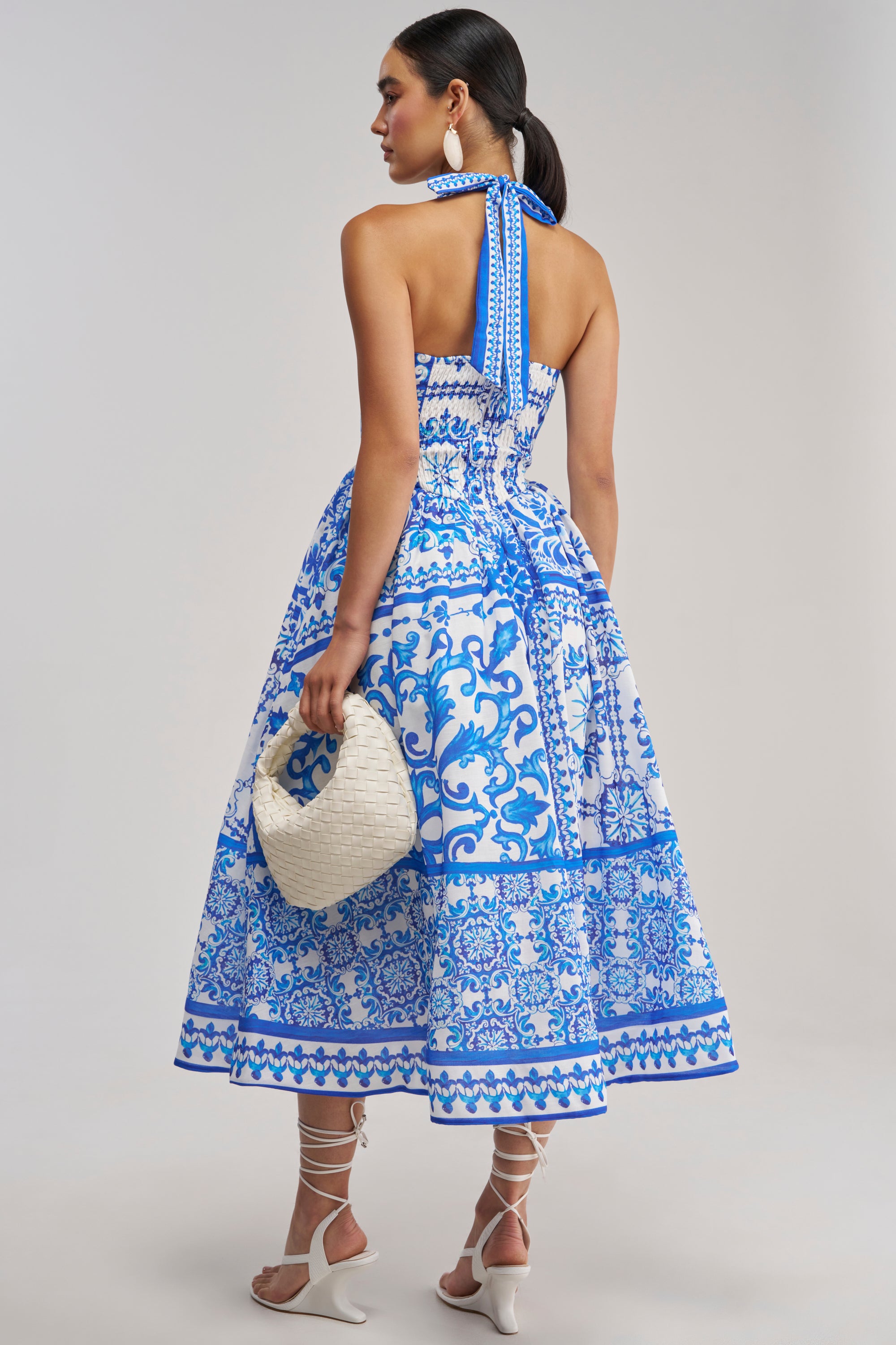 A woman wearing the COCO CRUSH PRINTED HALTER MIDI DRESS, styled with a white woven handbag, white strappy heels, and large round earrings, stands against a neutral background.