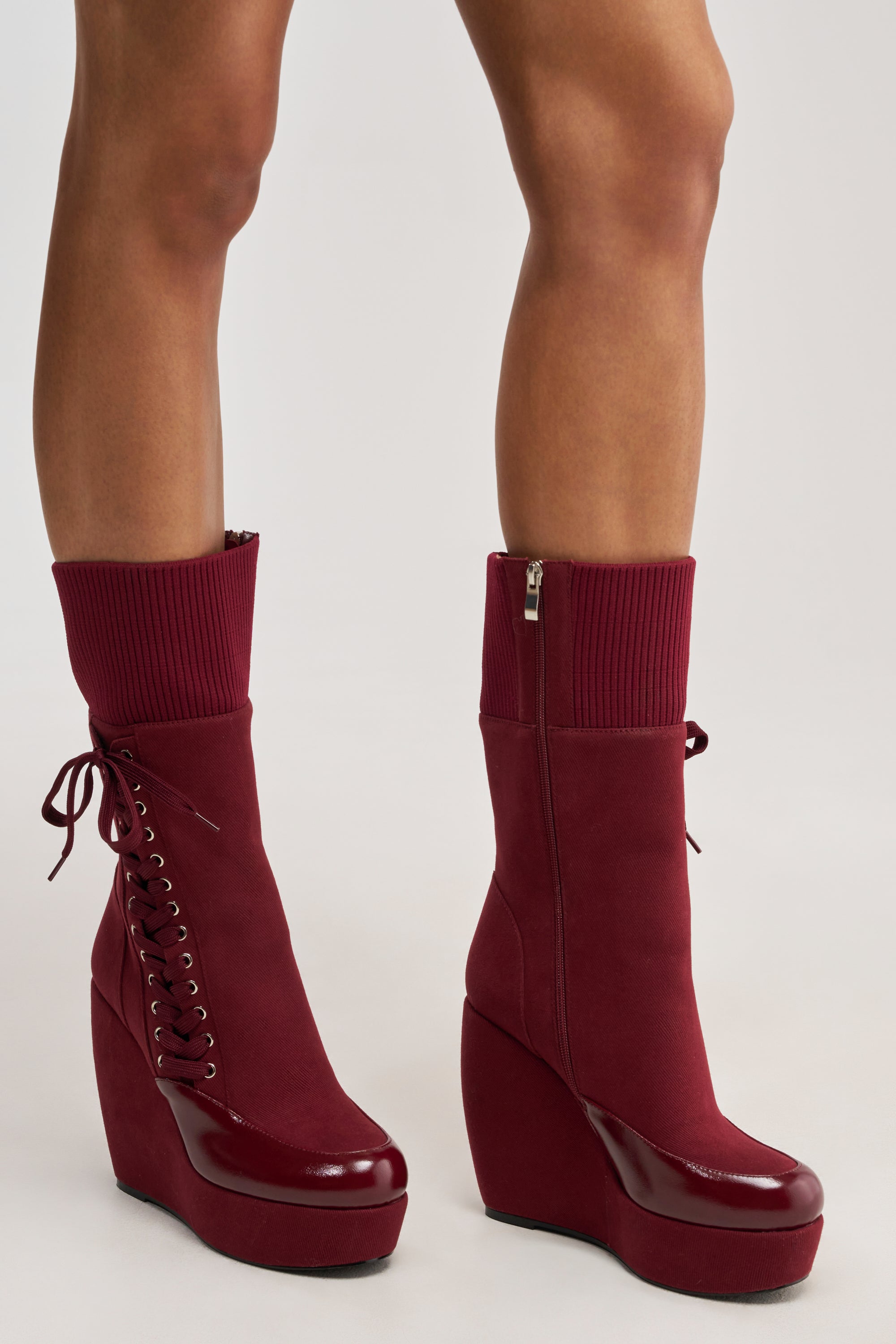 A person wearing AKIRA GOLD WHATEVER BURGUNDY CANVAS WEDGE BOOTIE—knee-high boots with wedge heels, side zippers, lace-up detail on one boot, ribbed knit cuffs, and glossy toe caps—stands on a light surface.