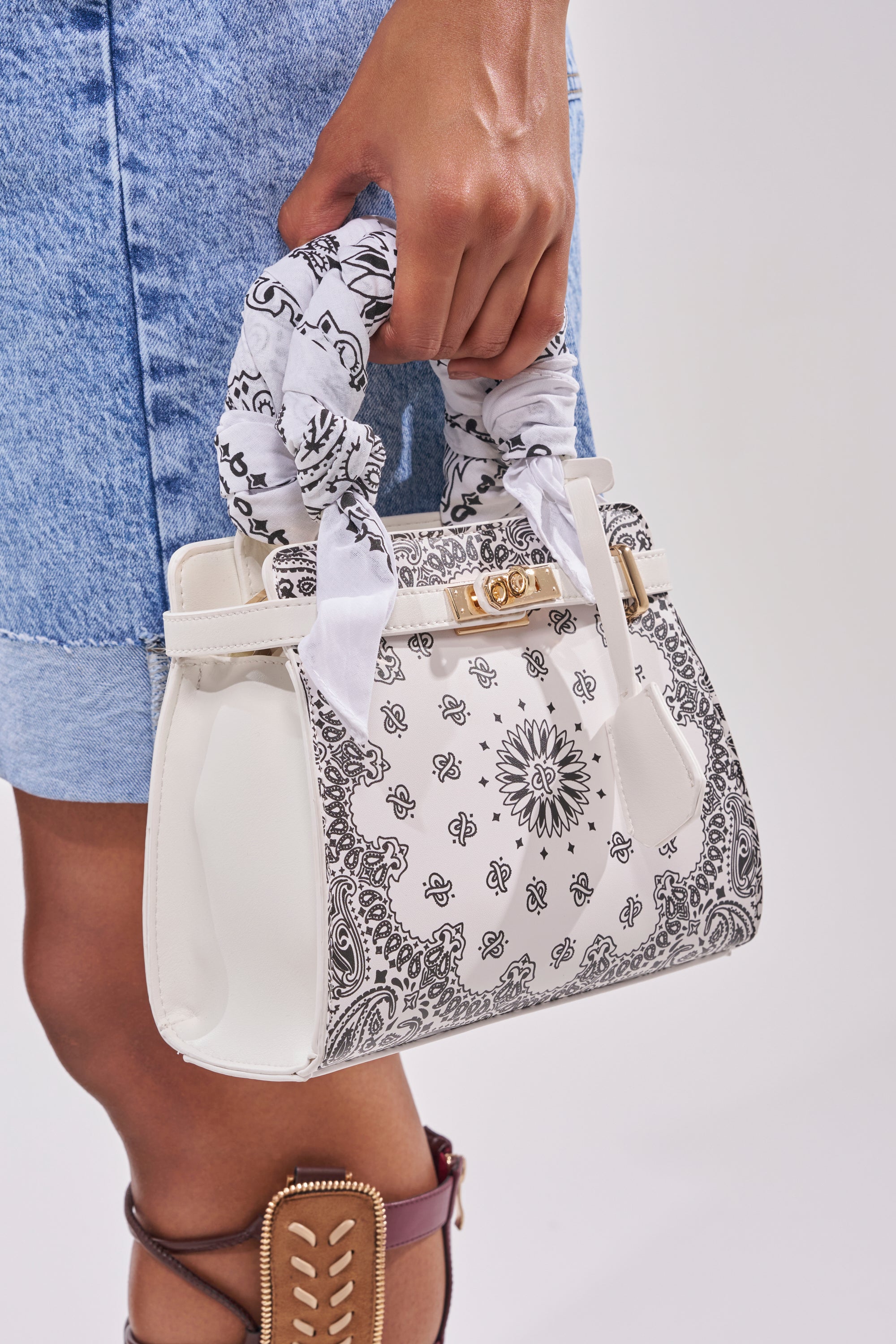 A person in a denim skirt holds the VIVIENNE PAISLEY PRINT BAG, featuring a handle wrapped in a black-and-white scarf. Their visible leg and sandal with tan accents enhance the western festival vibe. 3
