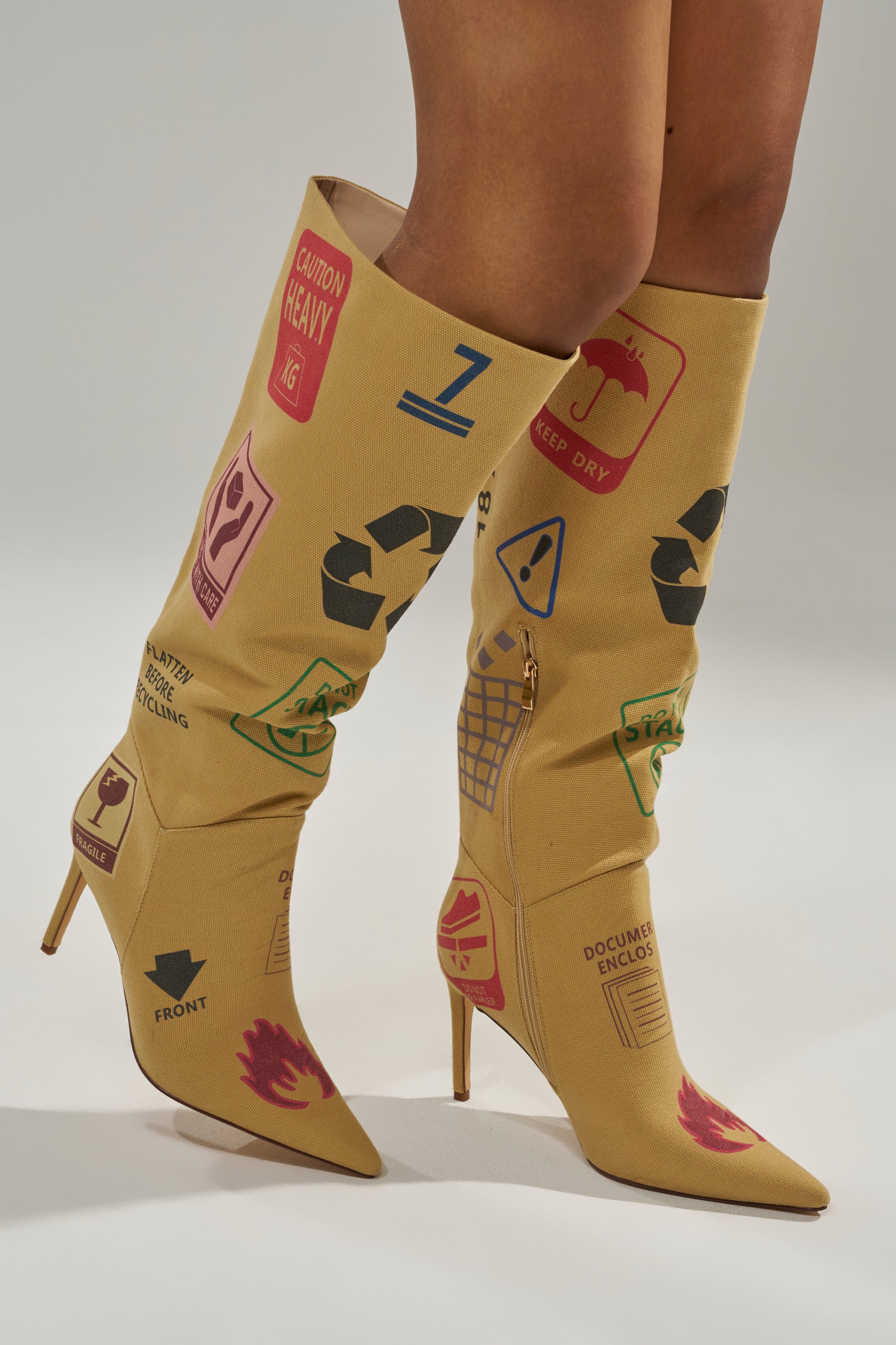 The AKIRA GOLD WHOLE PACKAGE TAN STILETTO BOOT, featuring a pointed toe and colorful shipping and recycling symbols like “Caution Heavy” and “Keep Dry,” is worn by a model standing on a neutral background. 1