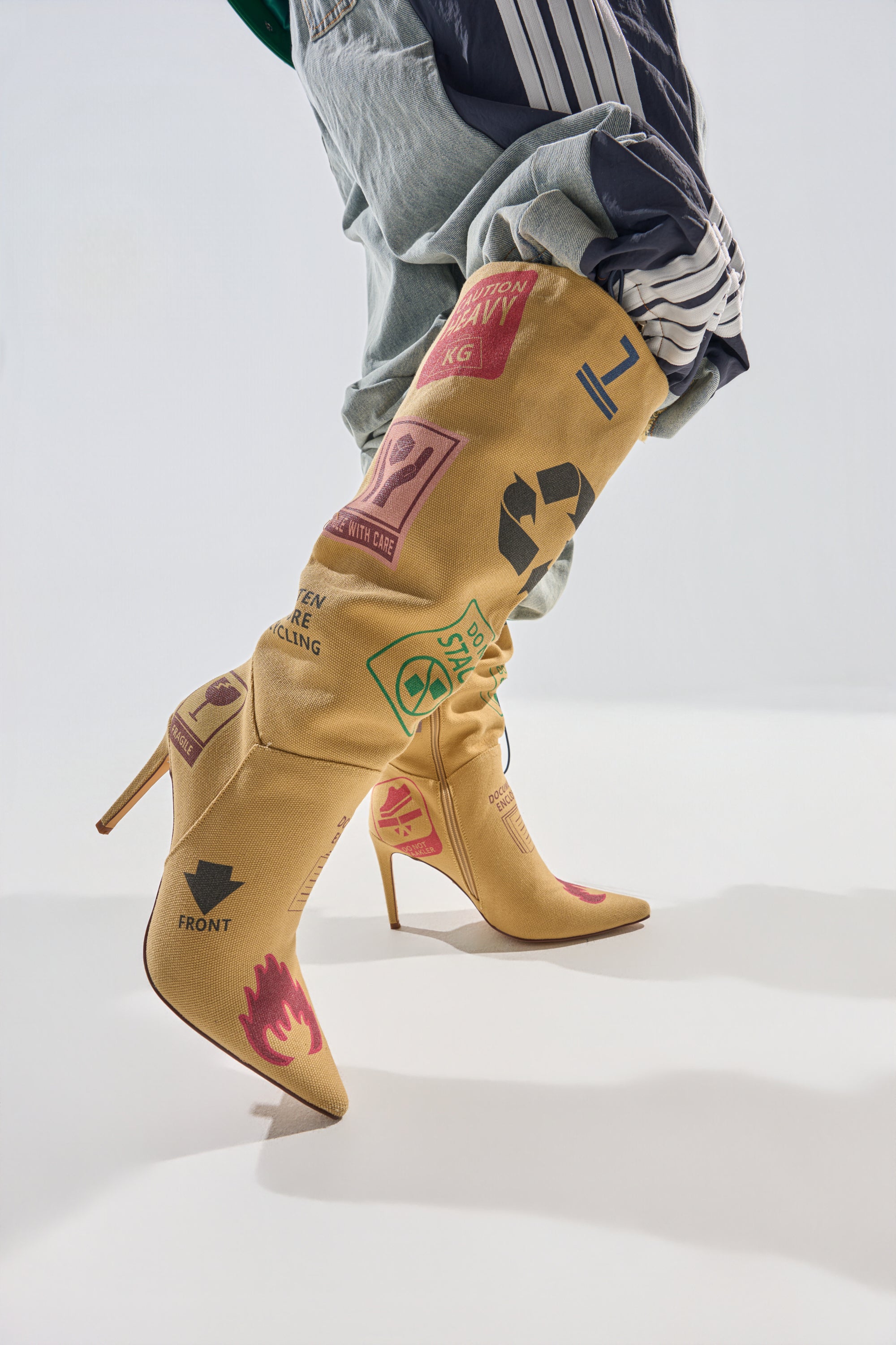Wearing the AKIRA GOLD WHOLE PACKAGE TAN STILETTO BOOT adorned with colorful symbols and recycling icons, a person pairs them with loose gray pants featuring white stripes, standing against a plain light background. 0