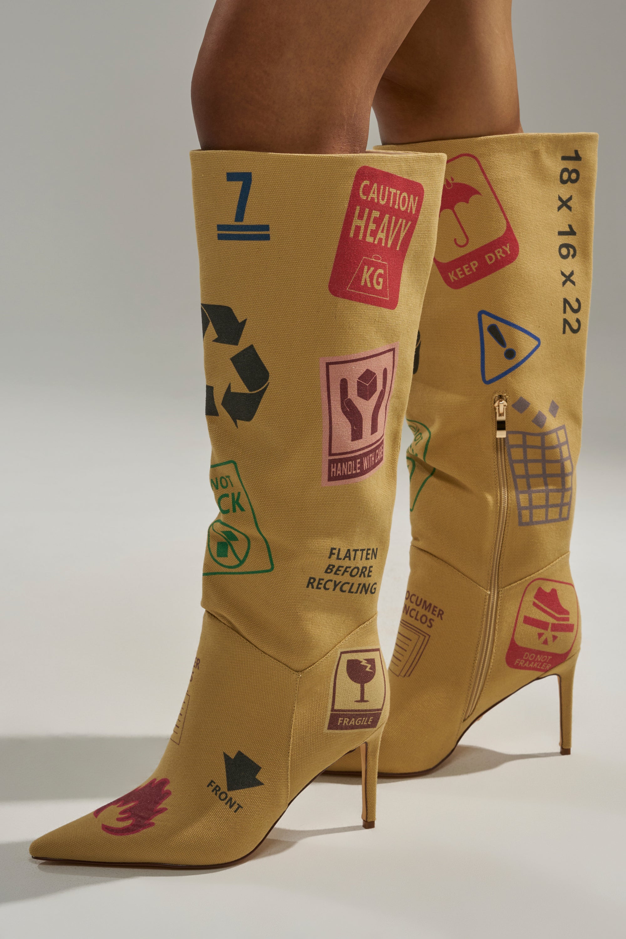 A person wears the AKIRA GOLD WHOLE PACKAGE TAN STILETTO BOOT, a pointed toe, knee-high boot in tan with bold shipping and recycling symbols like “Caution Heavy,” “Keep Dry,” and various package handling icons. 4